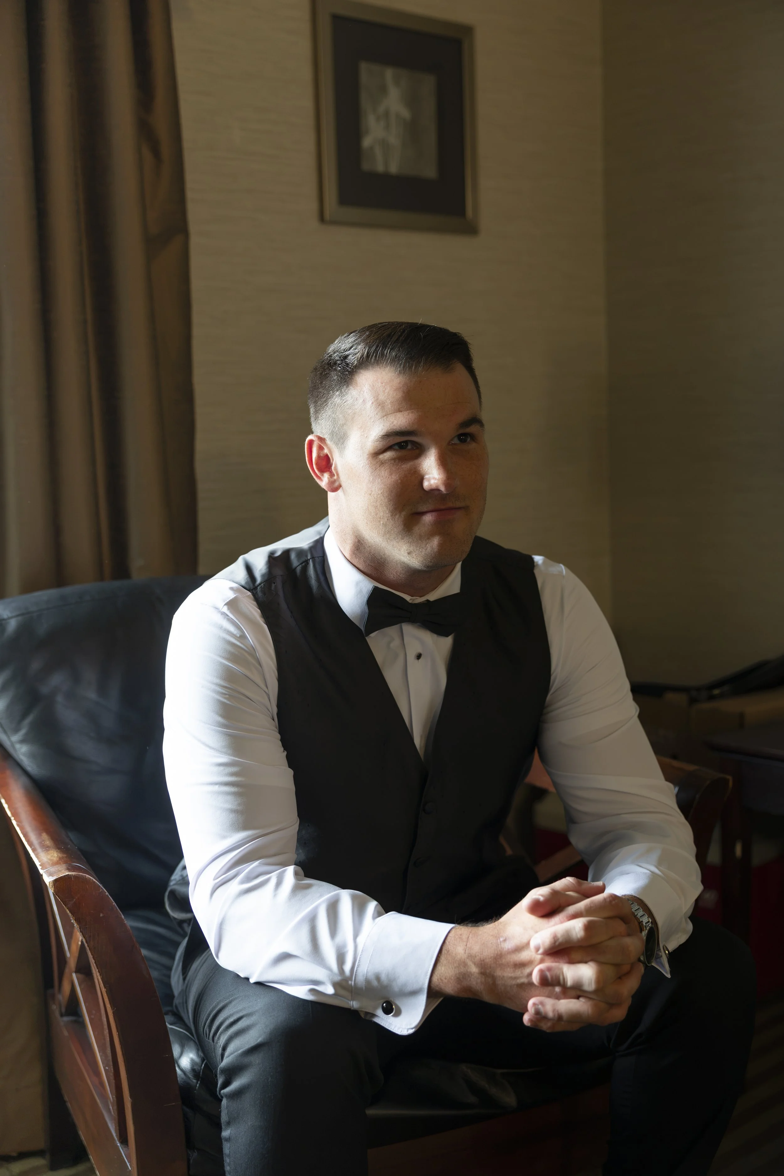A man in formal attire, including a white dress shirt, black vest, and bowtie, sitting in a leather chair with his hands clasped, in a room with beige walls and a framed picture hanging behind him.