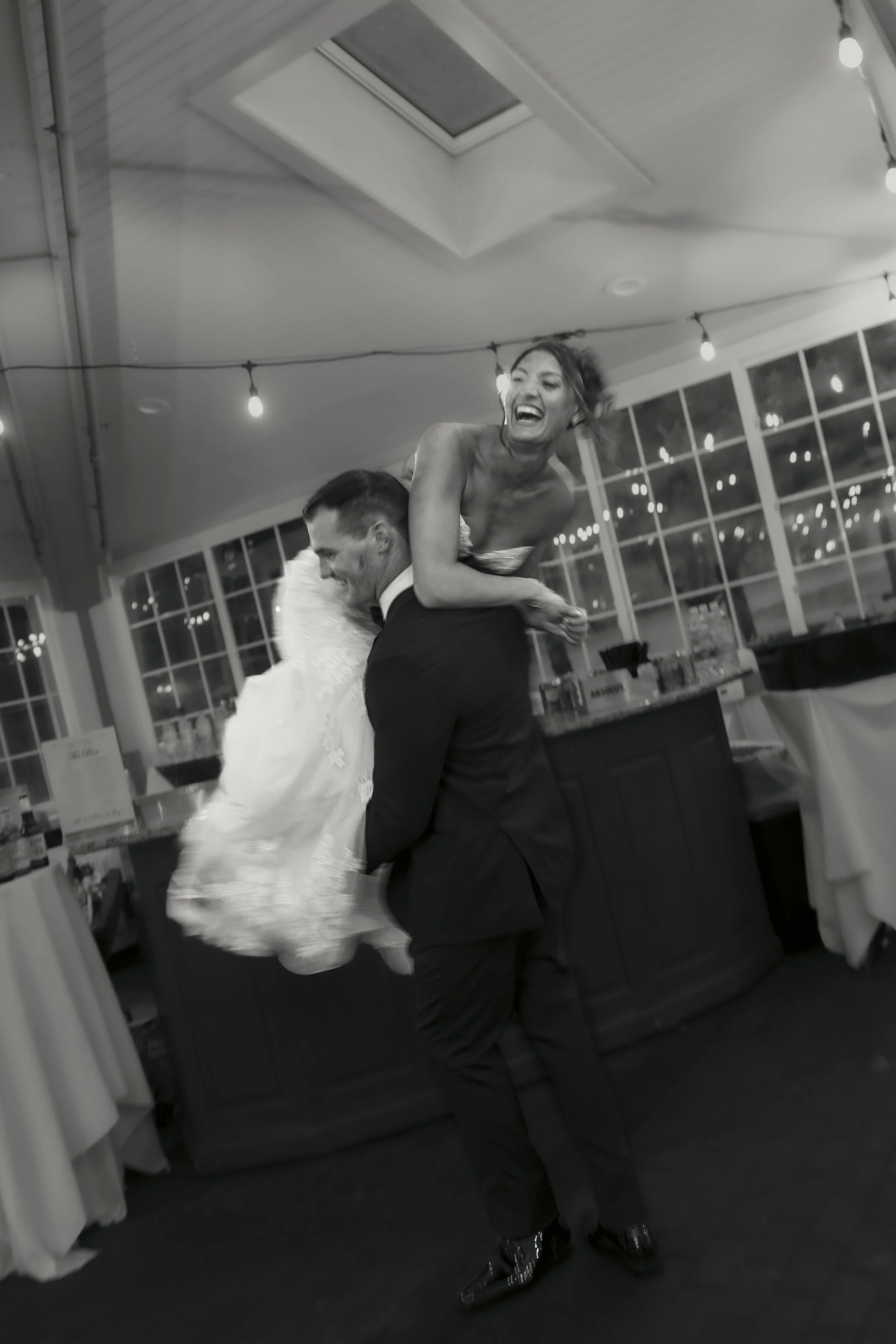 A joyful woman in a strapless dress being lifted by a man in a tuxedo at a celebration or wedding reception, with festive string lights and large windows in the background.