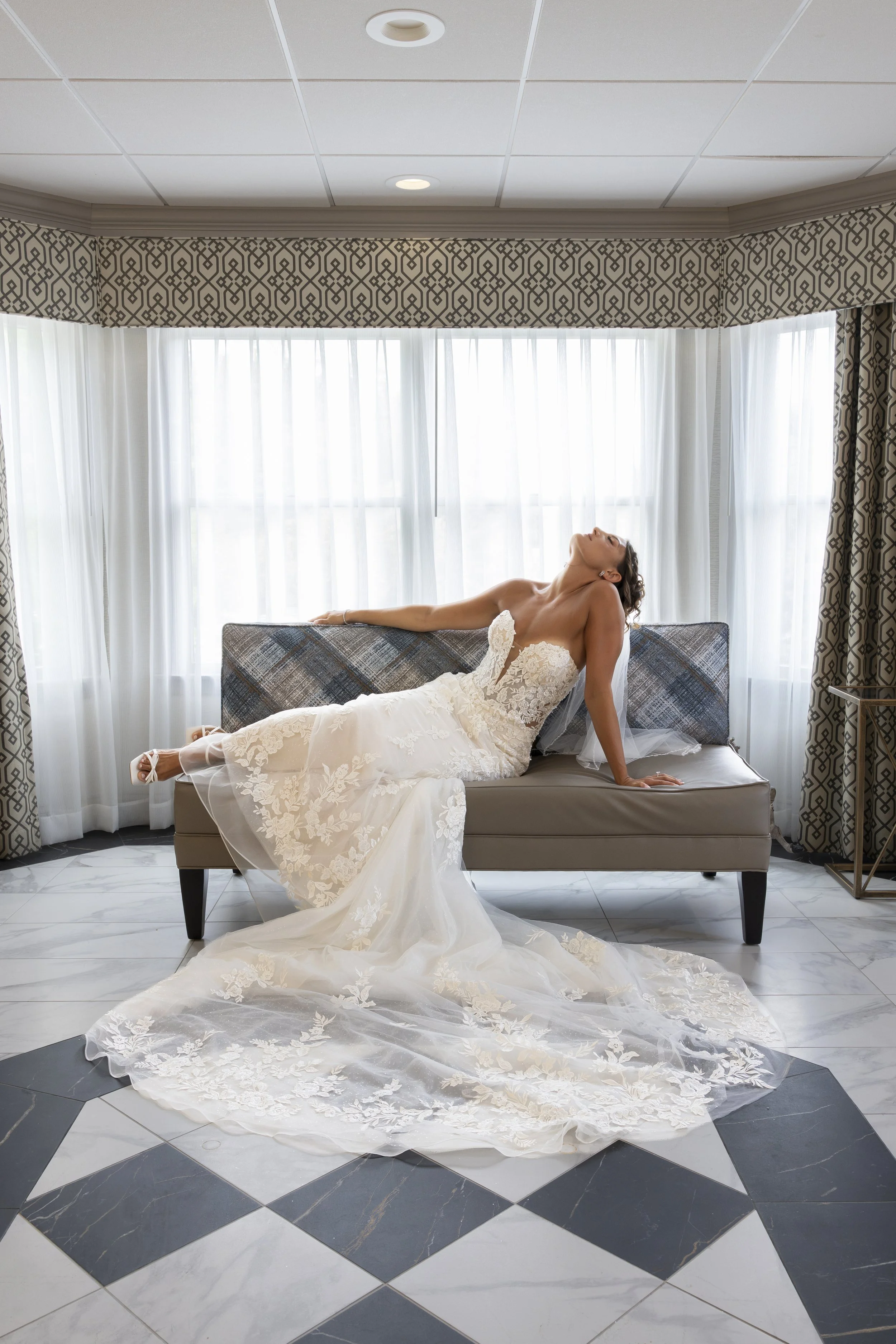 A bride in a white wedding dress sitting on a gray sofa with her head tilted back in a room with white curtains and patterned valances.