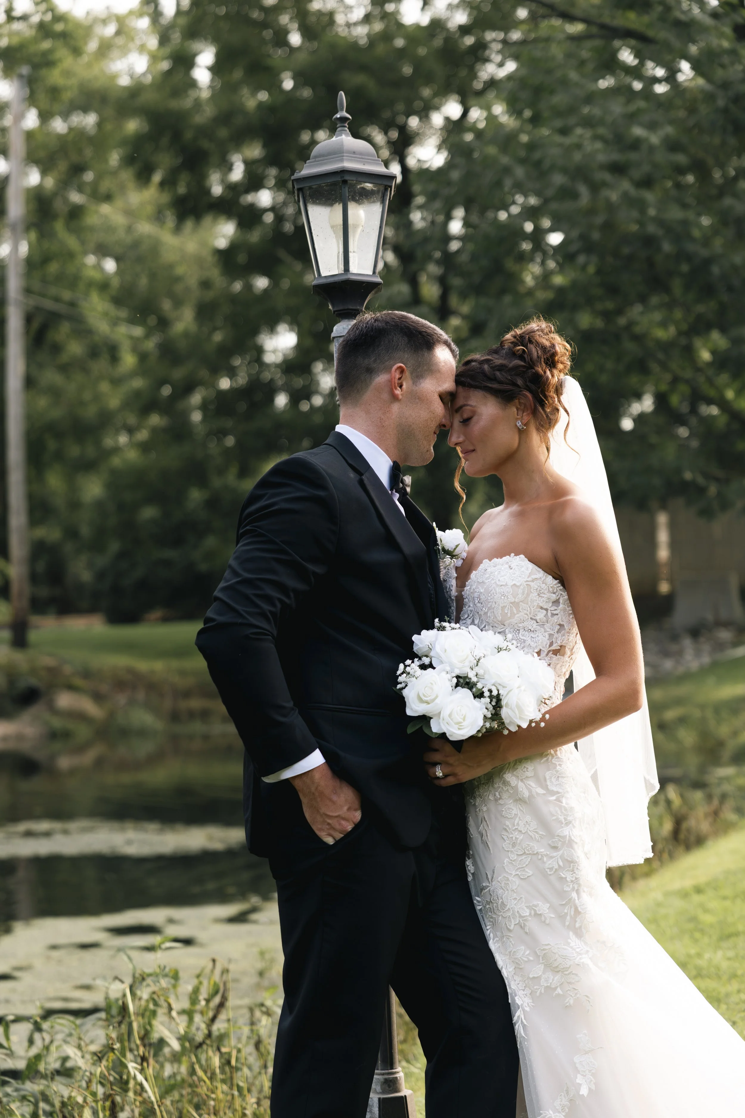 A bride and groom stand close together outdoors, touching foreheads with eyes closed, holding a bouquet of white roses, beside a vintage lamp post, with trees and a pond in the background.