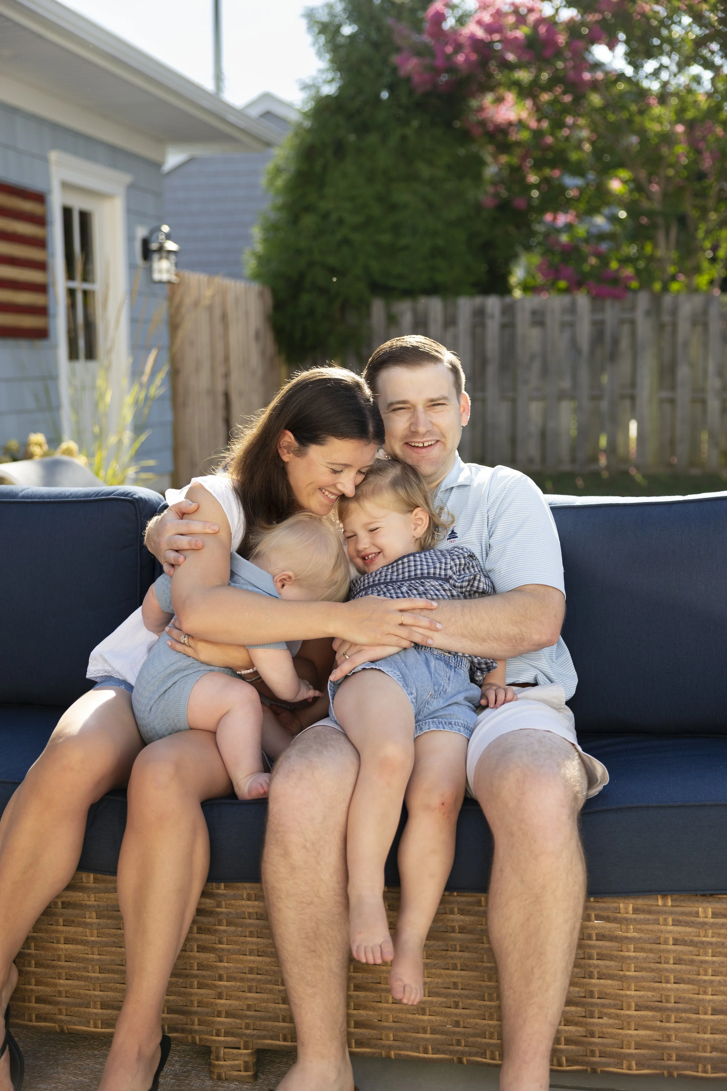 A happy family of five, sitting on a wicker outdoor sofa in a backyard, hugging and smiling. The father, mother, and three children are all close together. The backyard has a wooden fence, trees, and pink flowering bushes in the background.