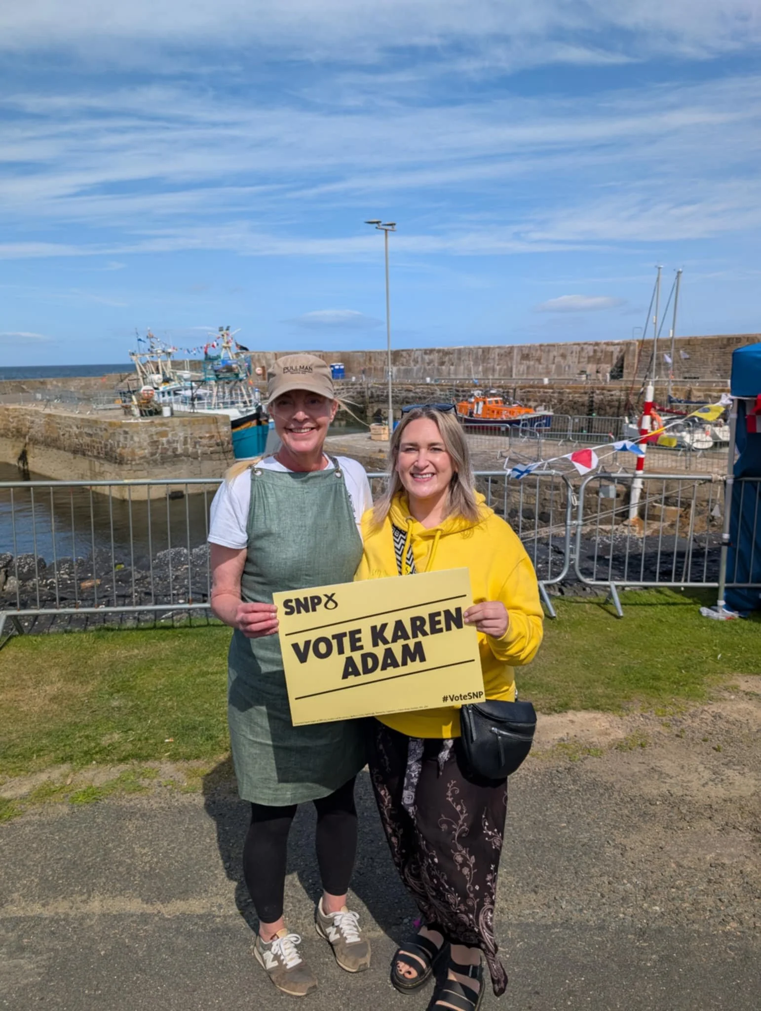 Two women standing outdoors near a harbor, holding a sign that reads 'Vote Karen Adam' for SNP, with boats and a stone pier in the background on a partly cloudy day.