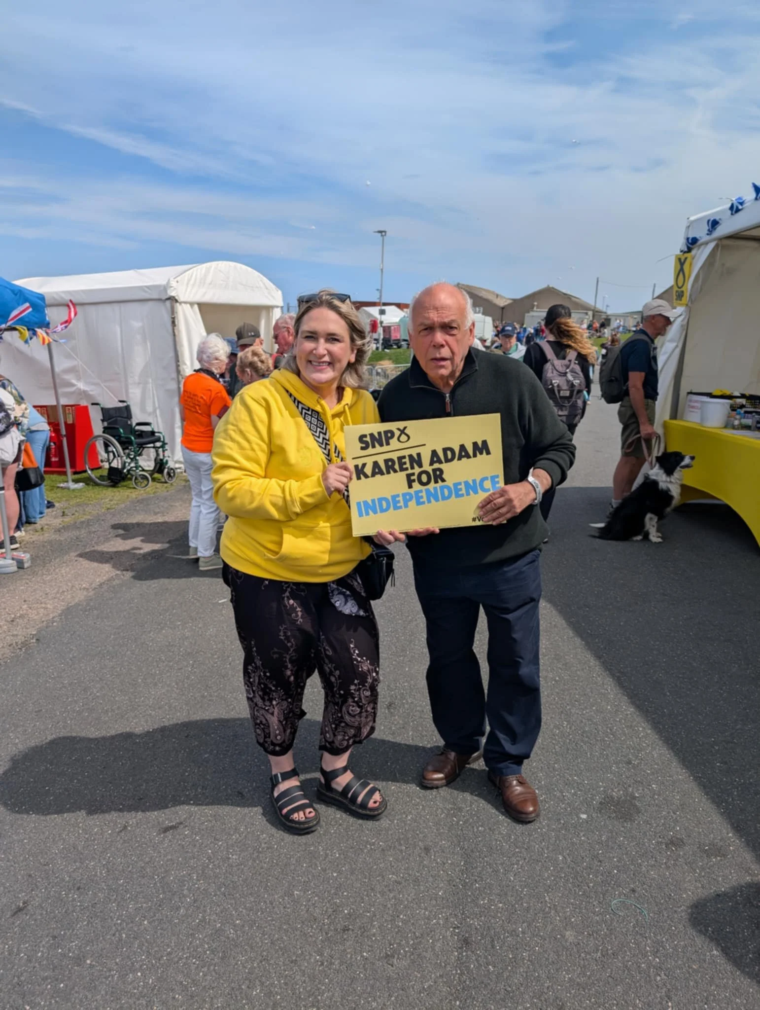 A woman and man at an outdoor event holding a sign that reads 'SNP & Karen Adam for Independence:' They are smiling and standing on a paved area with tents and people in the background.