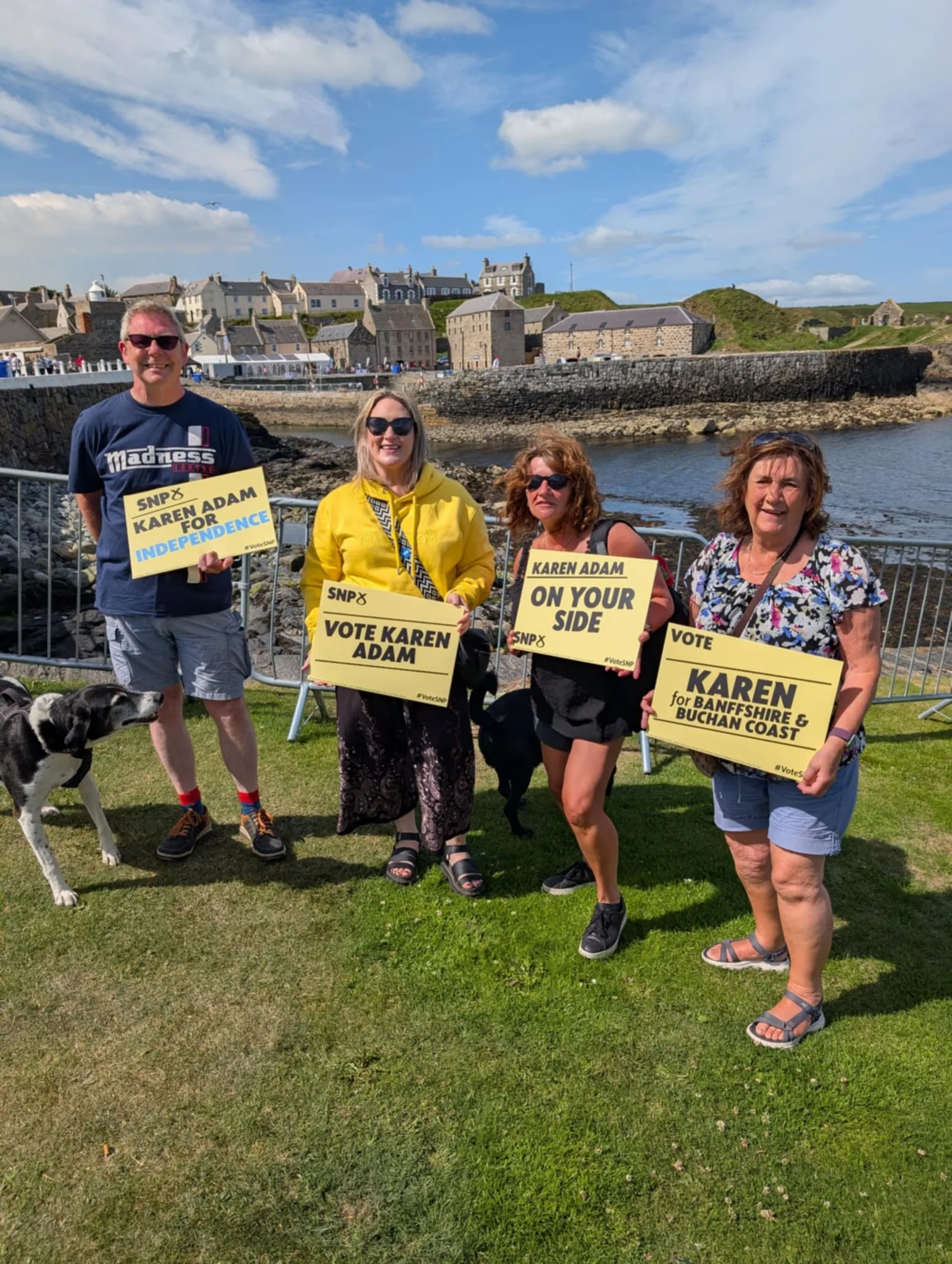 Four people holding campaign signs standing on a grassy area near a waterfront with historic buildings and a stone wall in the background. Two women and a man are wearing sunglasses, and there are dogs near them. The sky is partly cloudy.