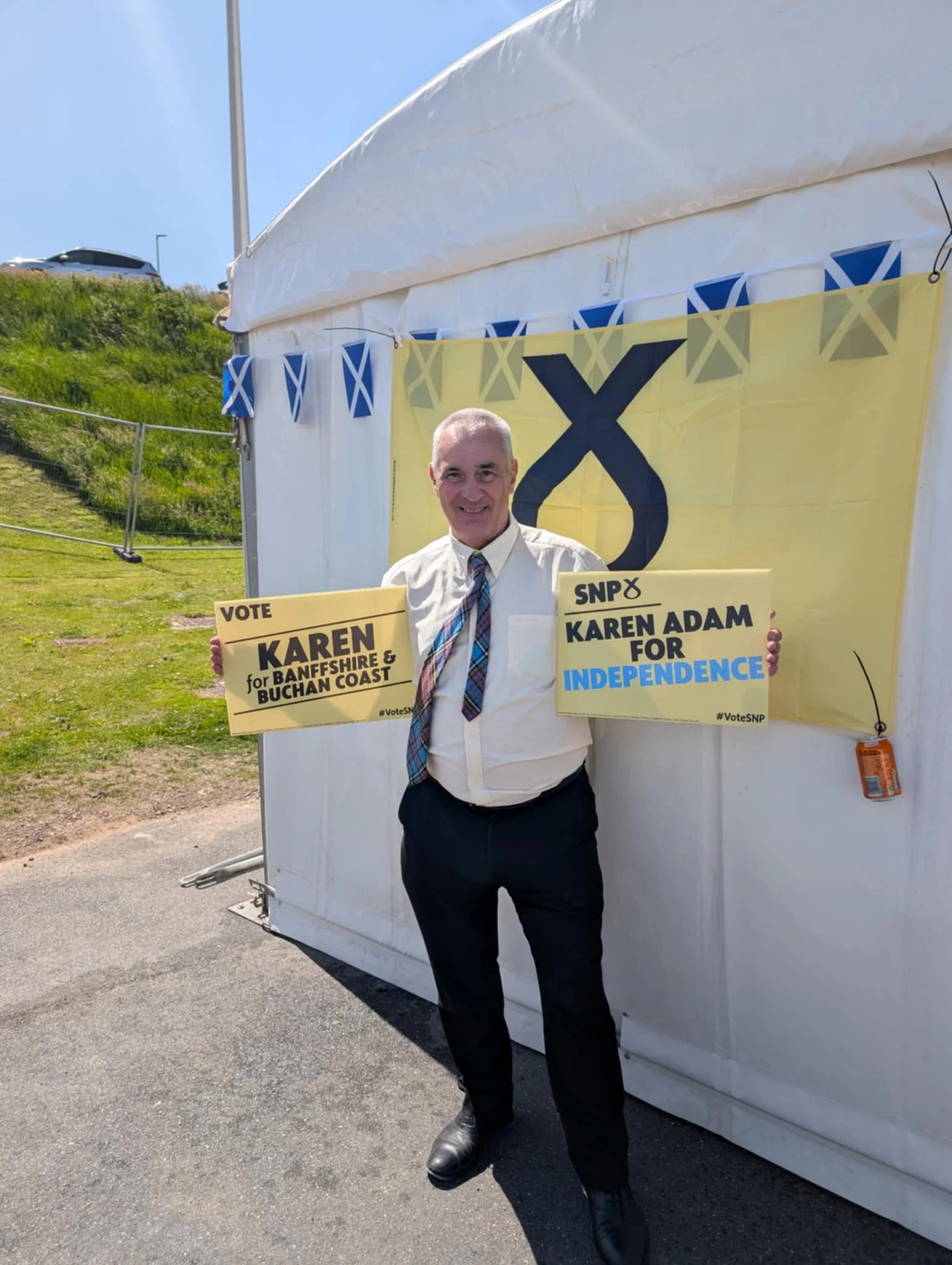 A man standing outdoors in front of a white tent, holding campaign signs for Karen Adam, with Scottish flags and a large banner with a Scottish saltire symbol behind him.