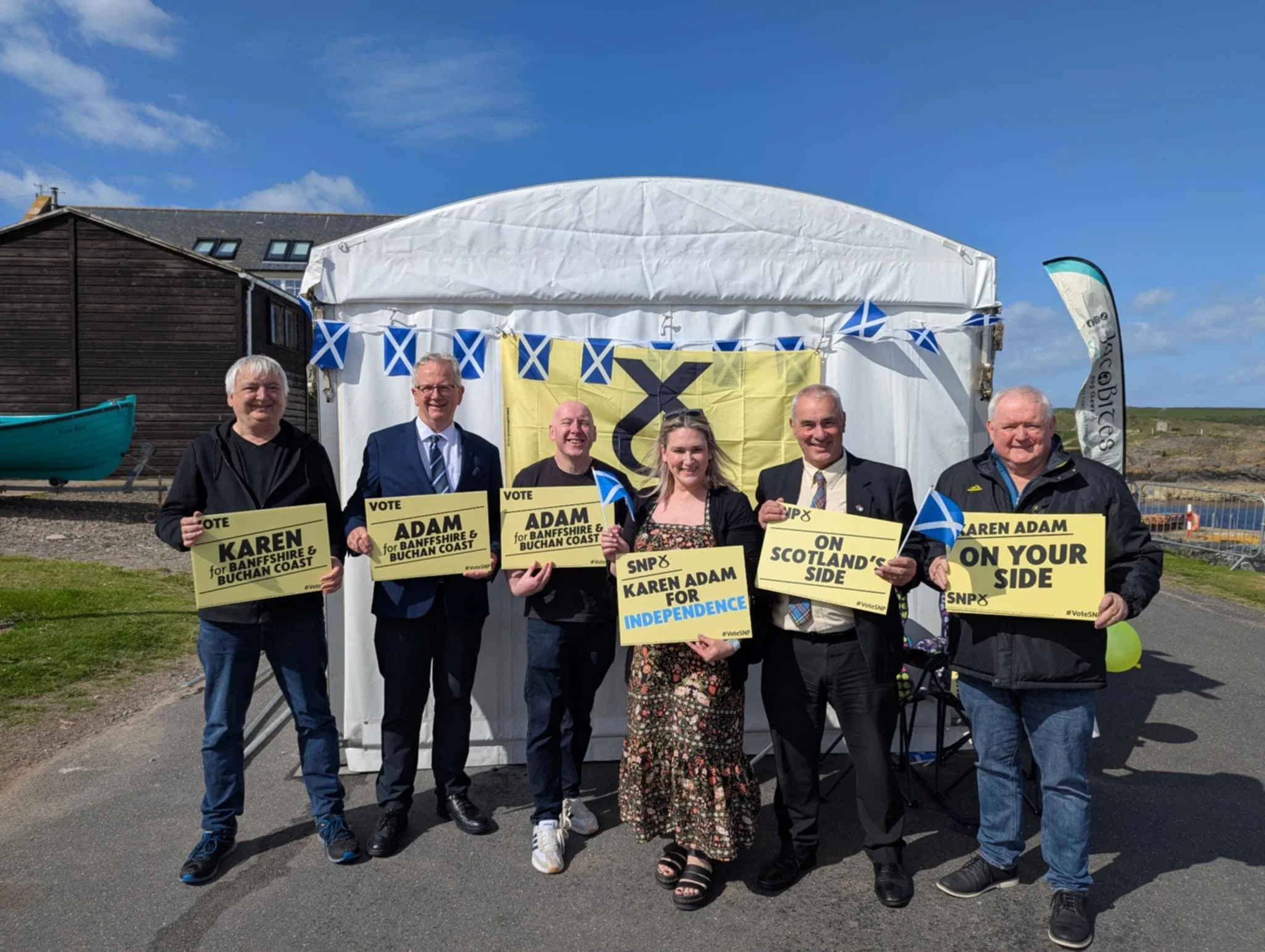 Group of six people standing outdoors, holding campaign signs and Scottish flags, with a blue sky and a small boat in background.