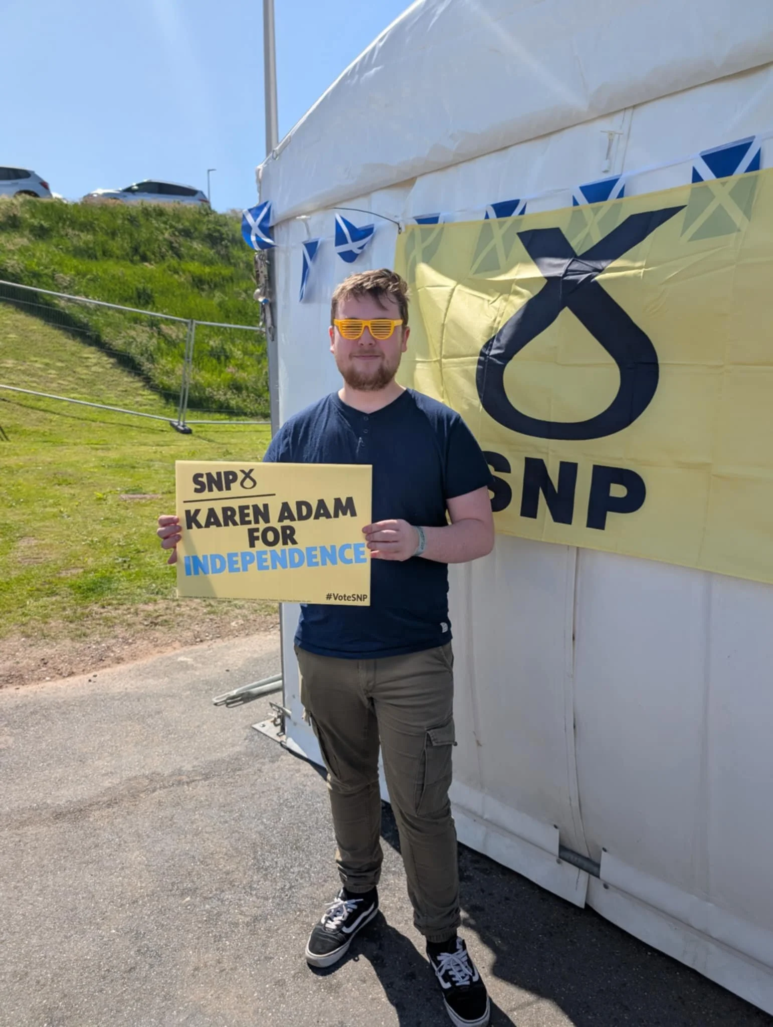 A man standing outdoors holding a yellow sign that says "SNPs KAREN ADAM FOR INDEPENDENCE" in front of a large yellow SNP banner on a white tent, with a grassy hill and parked cars visible in the background.