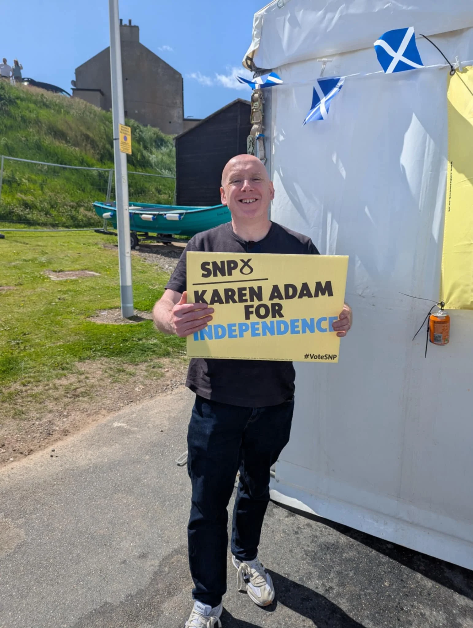 A man smiling and holding a campaign sign that reads "SNP KAREN ADAM FOR INDEPENDENCE" standing outdoors on a sunny day.