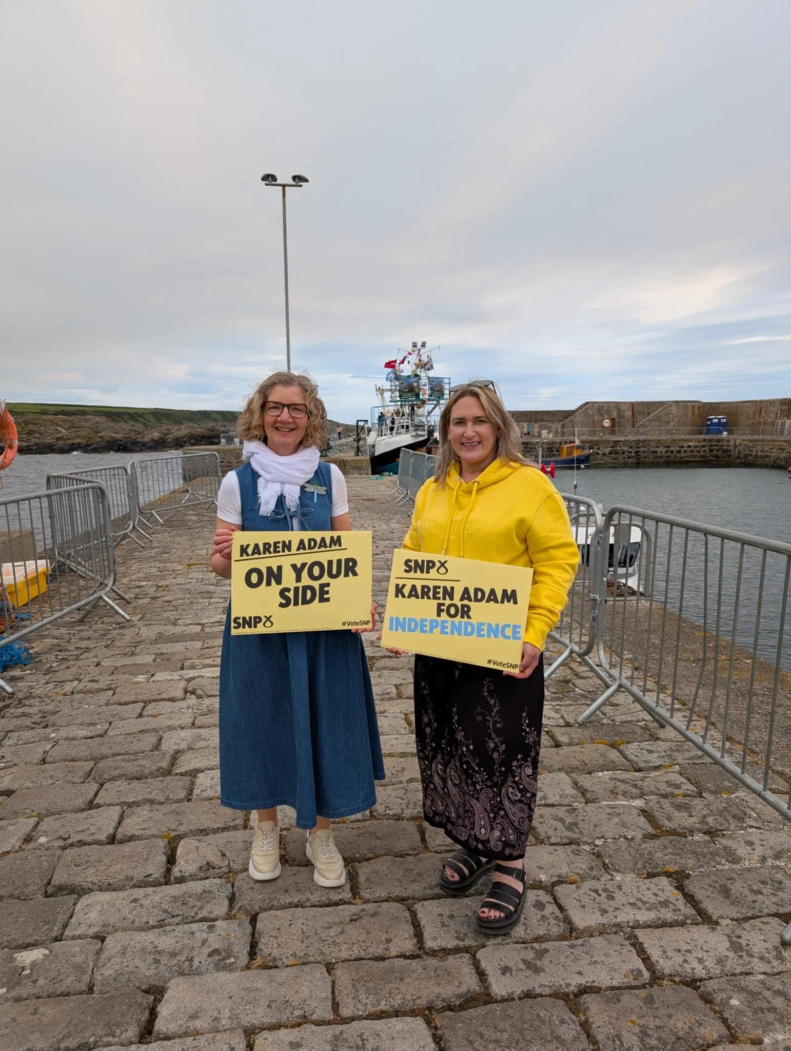 Two women standing on a stone dock by the water, holding signs supporting Karen Adam and independence, with a boat and cloudy sky in the background.