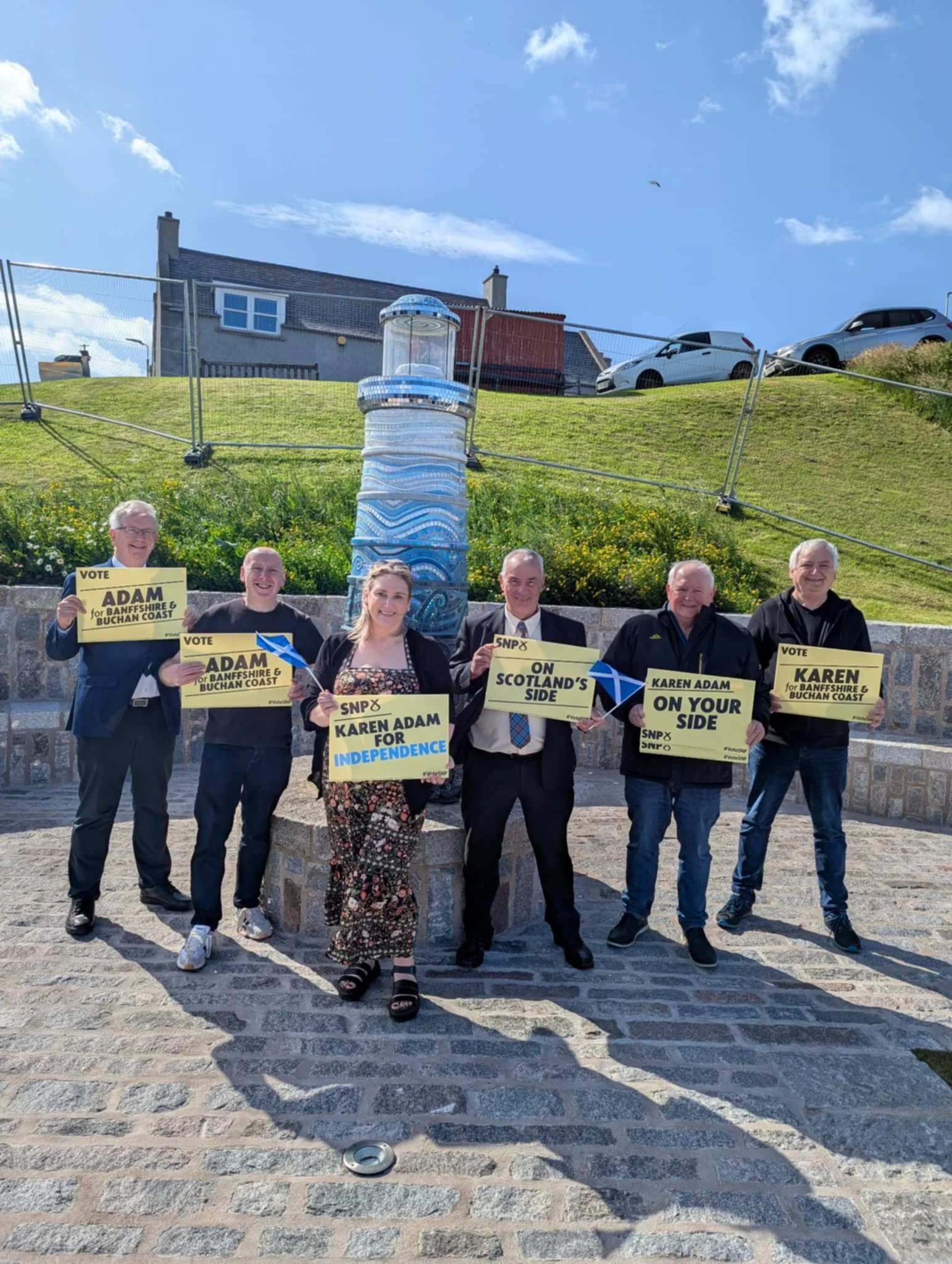 Six people standing outdoors holding political signs supporting Scottish independence, with a water fountain, a grassy hillside, and parked cars in the background.