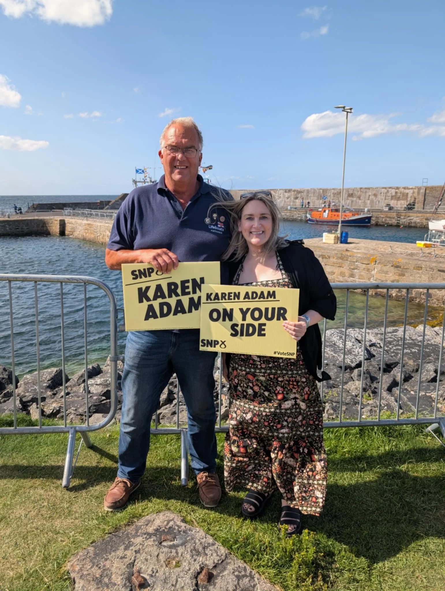 A man and woman standing by a seaside railing, holding campaign signs for Karen Adam, with the water and boats in the background on a sunny day.