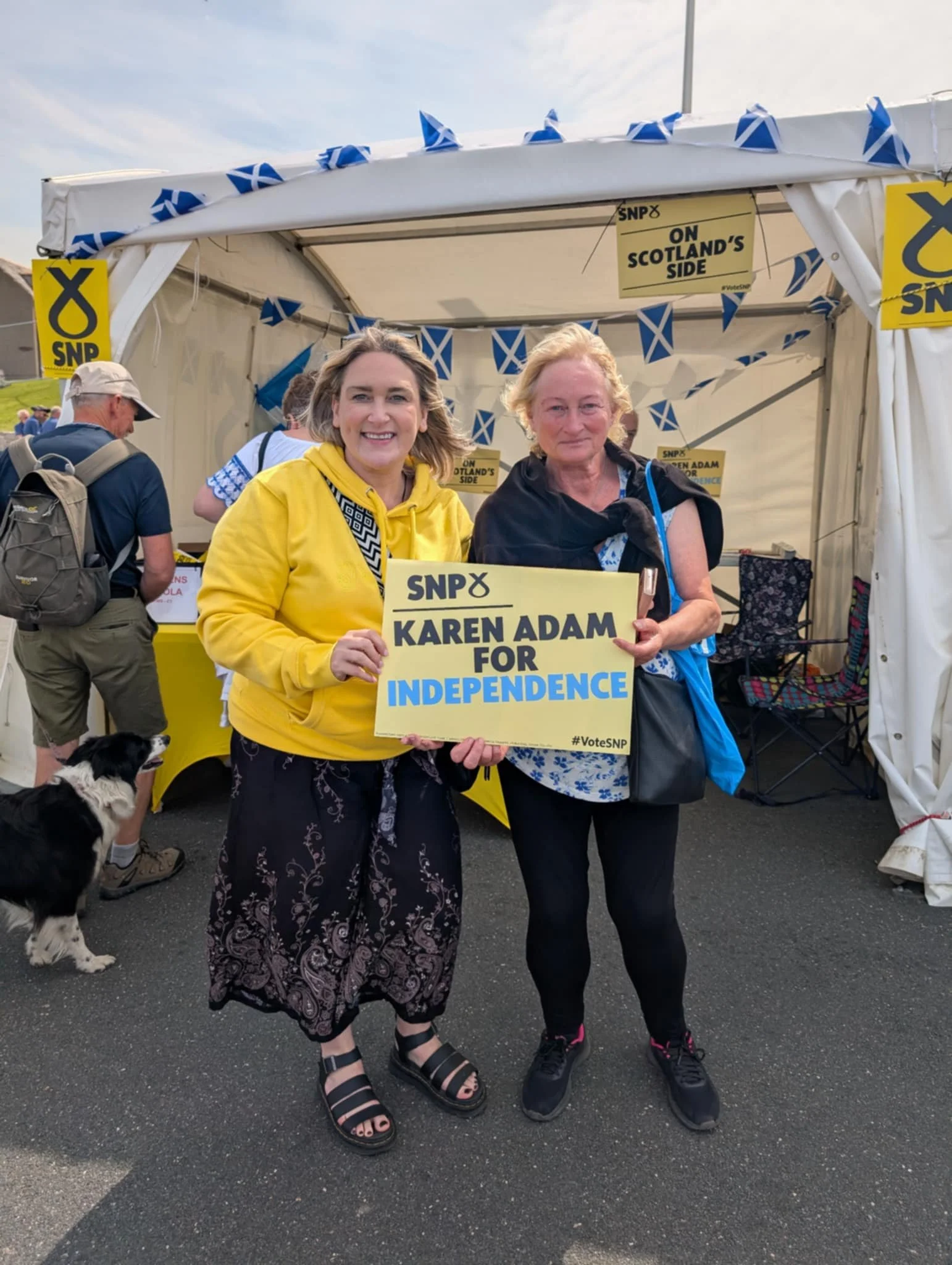 Two women holding a sign that says "Karen Adam for Independence" at a political event with a tent and Scottish flags in the background.
