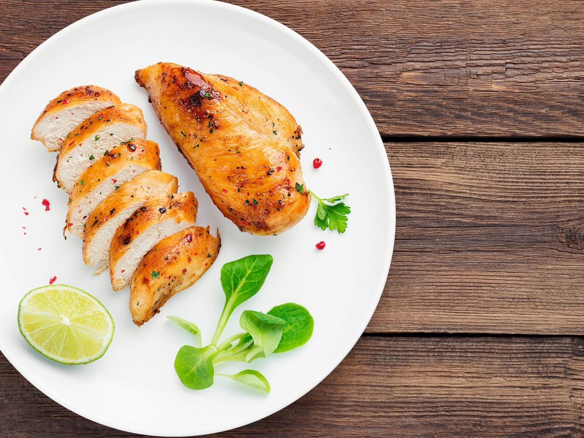 Grilled sliced chicken breast with lemon wedge and greens on white plate on wooden table.