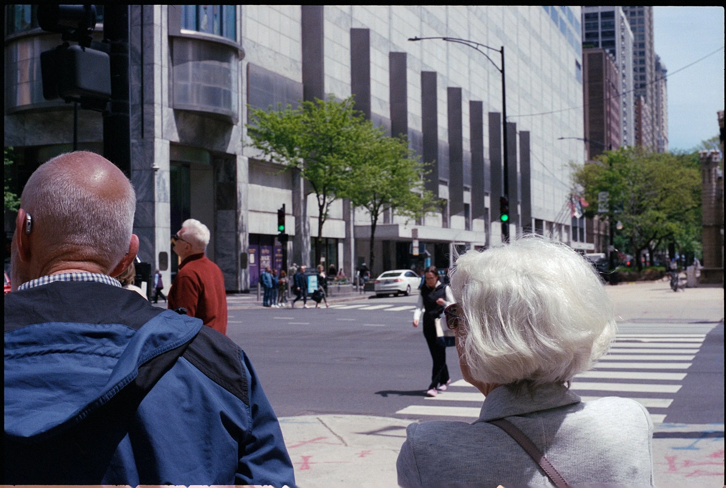 Elderly man and woman waiting to cross a city street at a pedestrian crosswalk, with modern buildings and trees in the background.