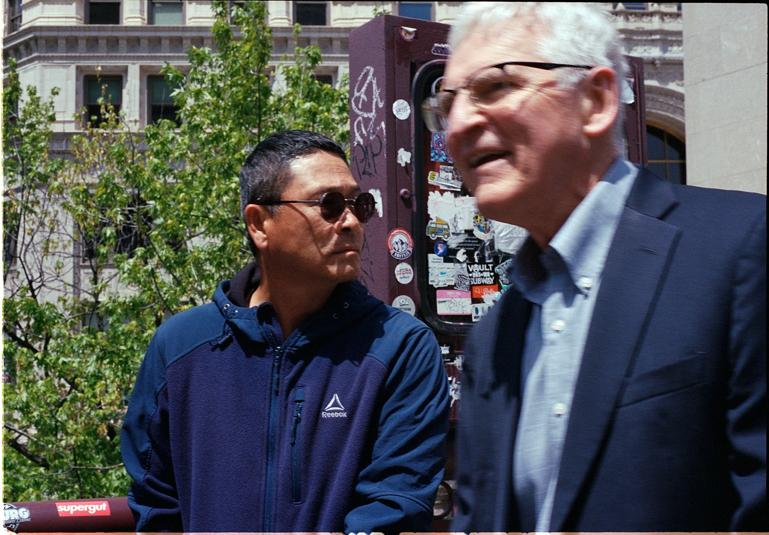 Two men engaged in conversation outdoors with trees and a sticker-covered utility box in the background.