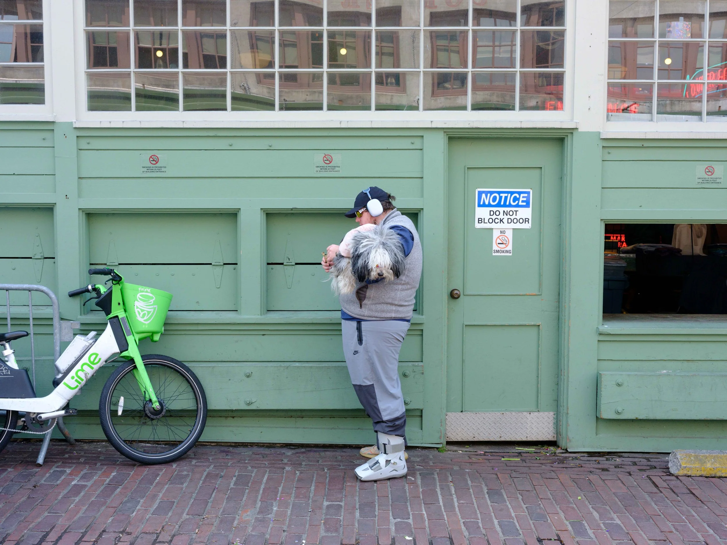 A man standing on a brick sidewalk holding a large dog in front of a green building with a sign that says "NOTICE: DO NOT BLOCK DOOR." The man is wearing a blue cap, sunglasses, ear protection, a gray vest, gray pants, and white boots. A lime green L