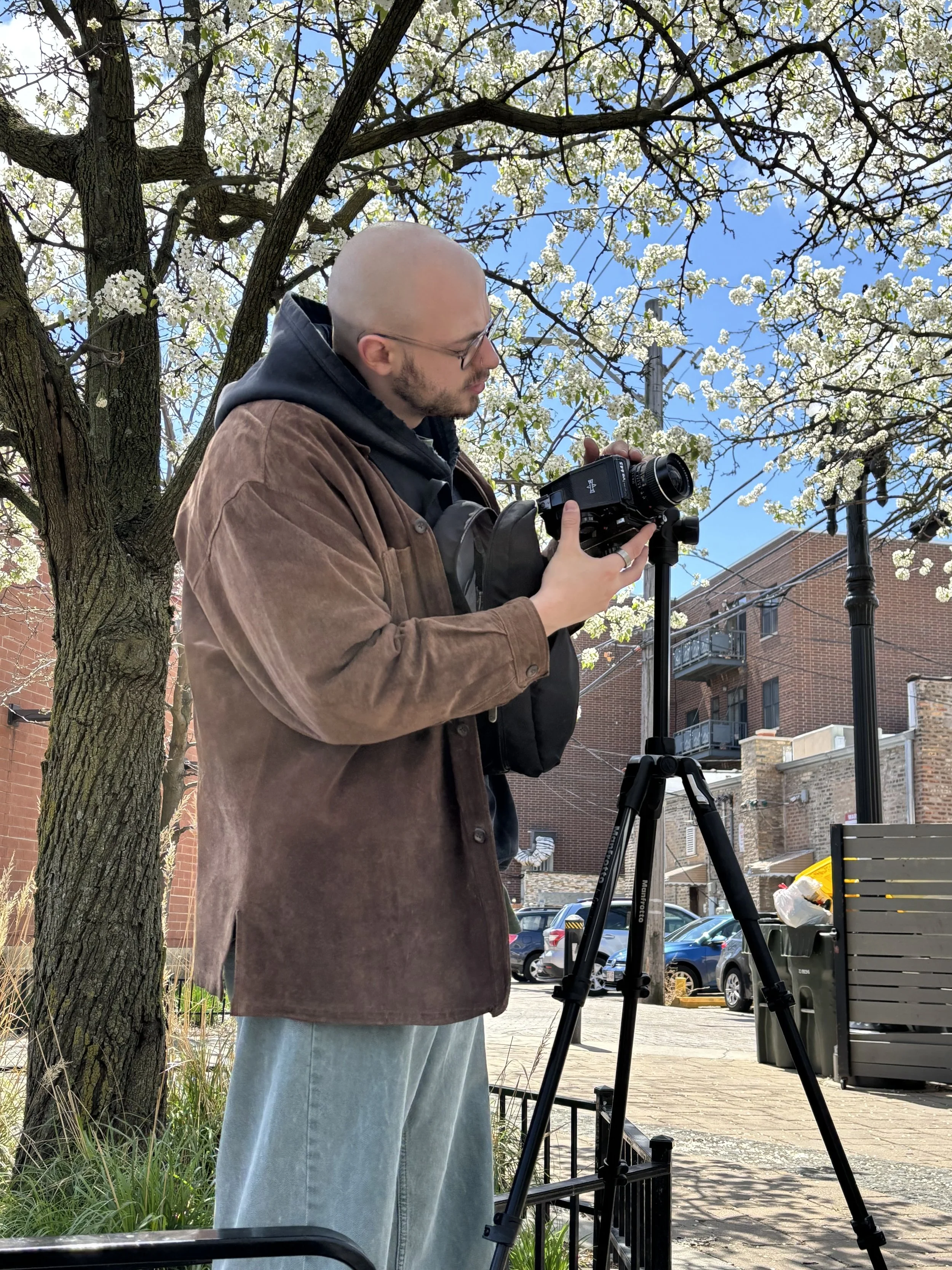 A man with a shaved head and glasses is looking at a camera mounted on a tripod outdoors. He is wearing a brown jacket over a hoodie. In the background, there are flowering trees, parked cars, a brick building, and a trash bin.