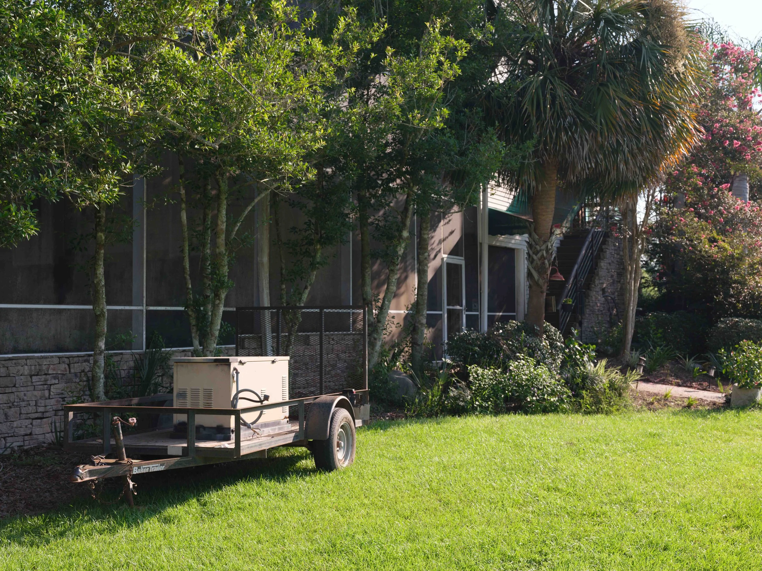 A yard with green grass, trees, bushes, and a house in the background. There is a small trailer with a box on it parked on the grass.
