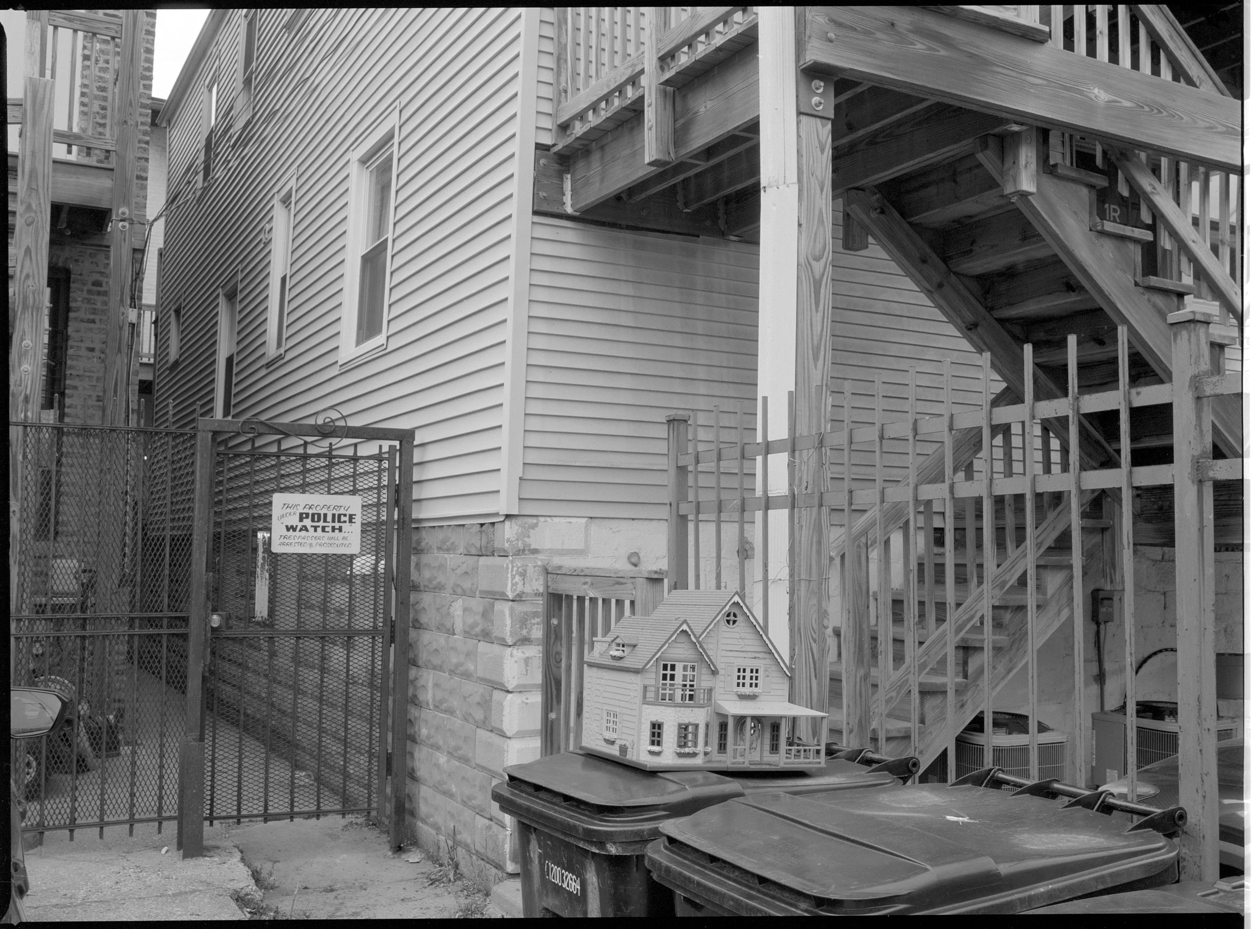 Backyard scene with a gated area on the left, a house with vinyl siding, and a wooden staircase on the right leading to an upper level. In the foreground, a plastic dollhouse is placed on top of three large trash bins, with a small window and porch v