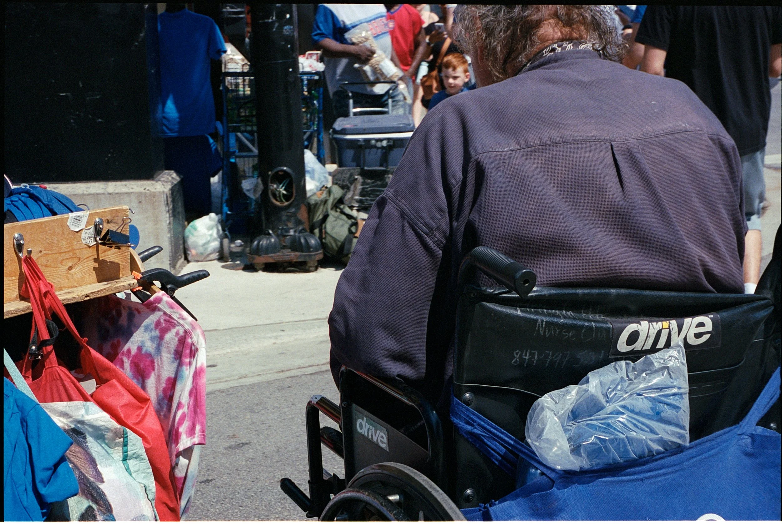 A person in a wheelchair, wearing a dark purple jacket, is sitting in front of a sidewalk sale with various clothes and bags on display. There are people and shopping carts in the background.
