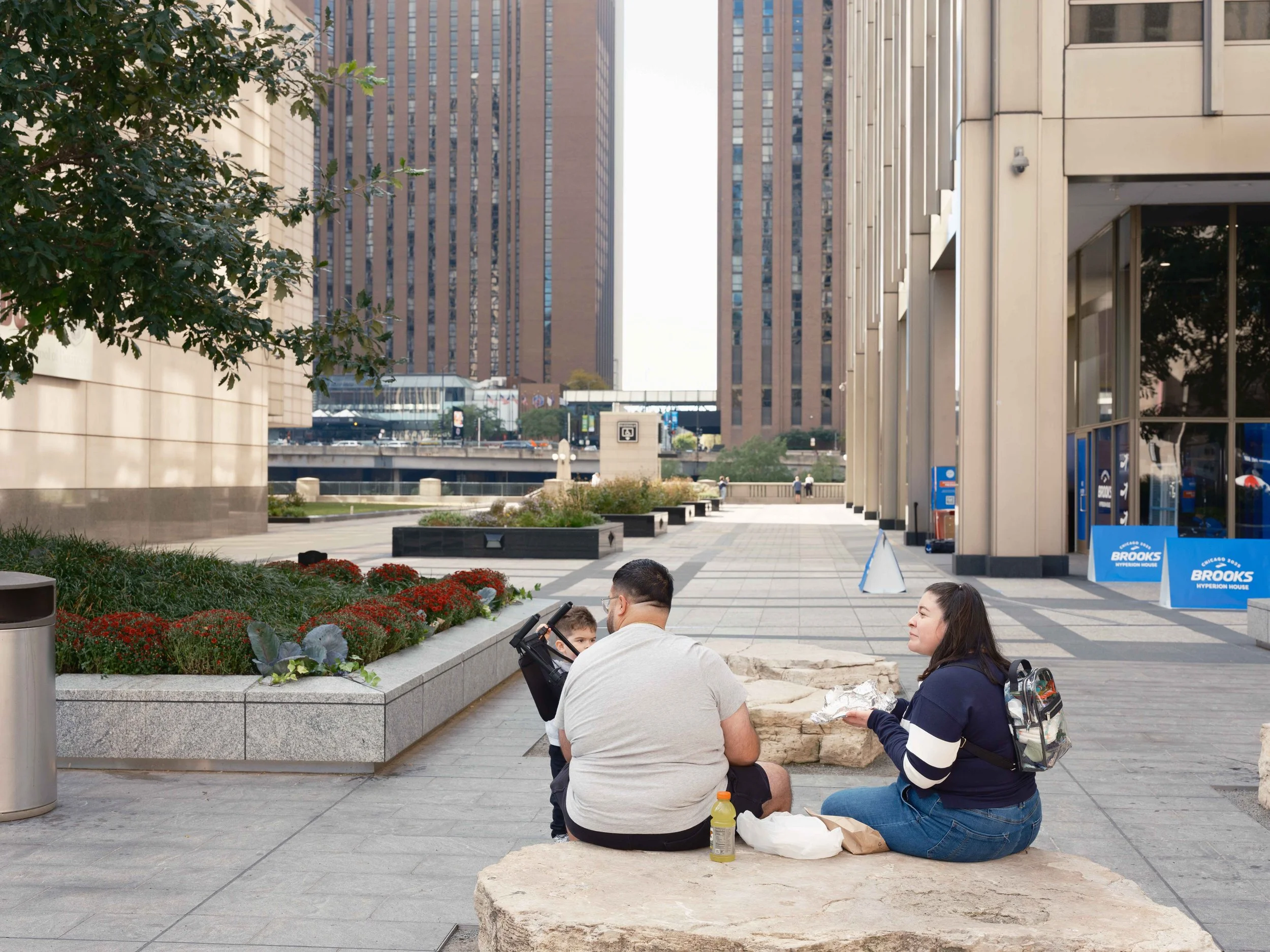 A group of people sitting on a stone bench in an urban park area, with tall buildings in the background. The scene includes a woman eating, a man, and a child.