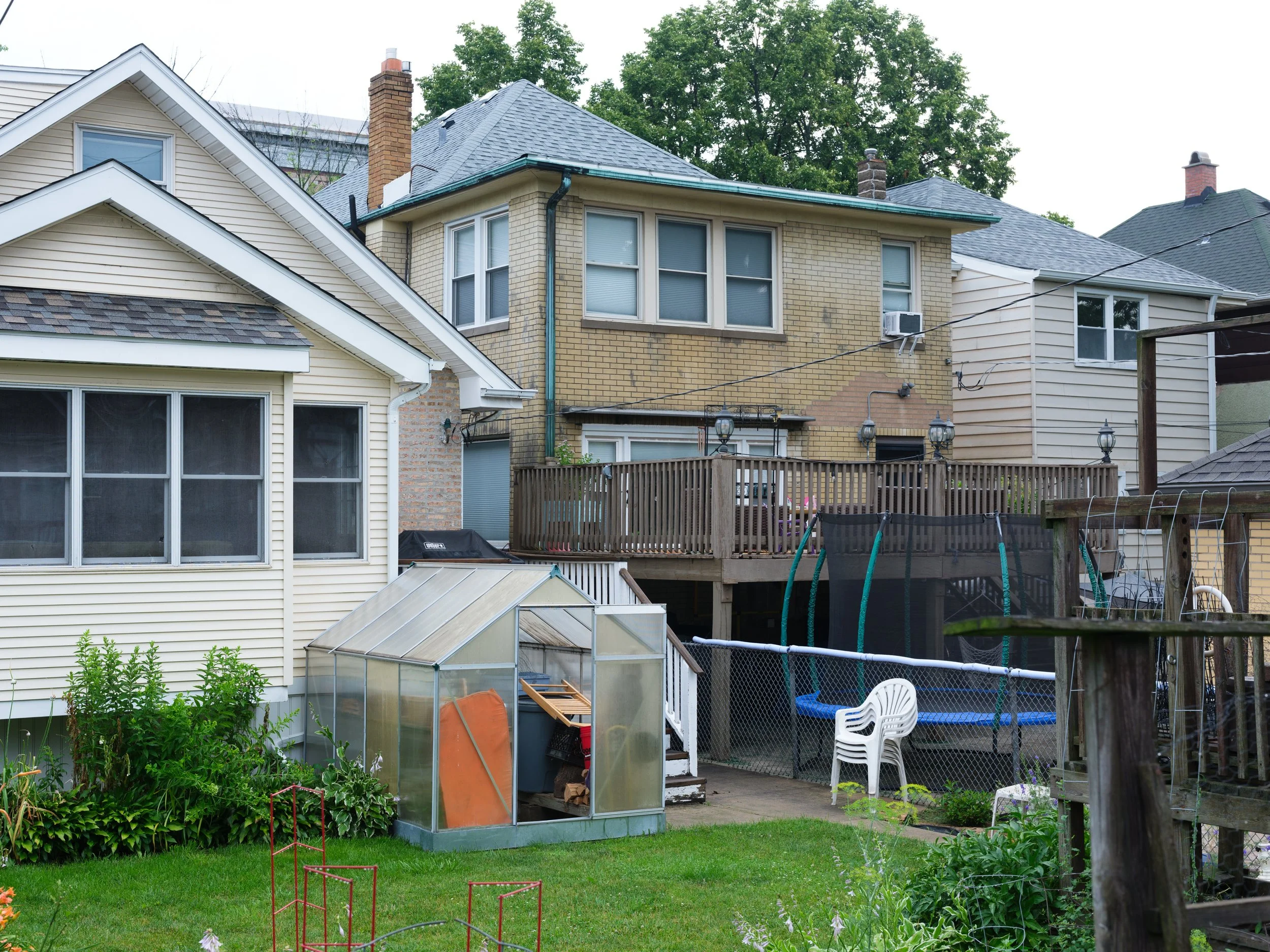 Backyard of a house with a greenhouse, a trampoline, a deck with outdoor furniture, and a small grassy area with plants.