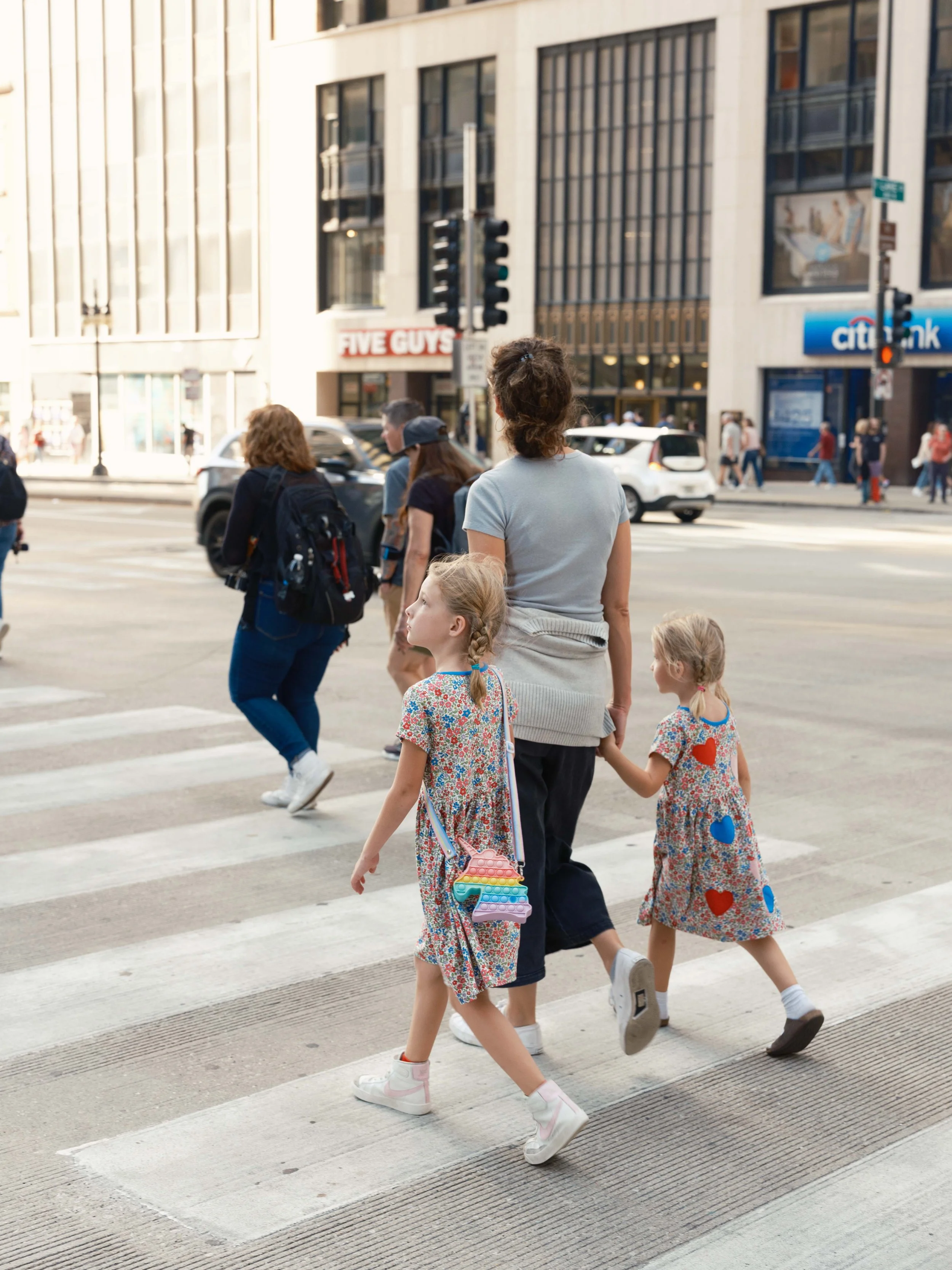 A woman walking with two young girls across a city crosswalk, with cars and buildings in the background.