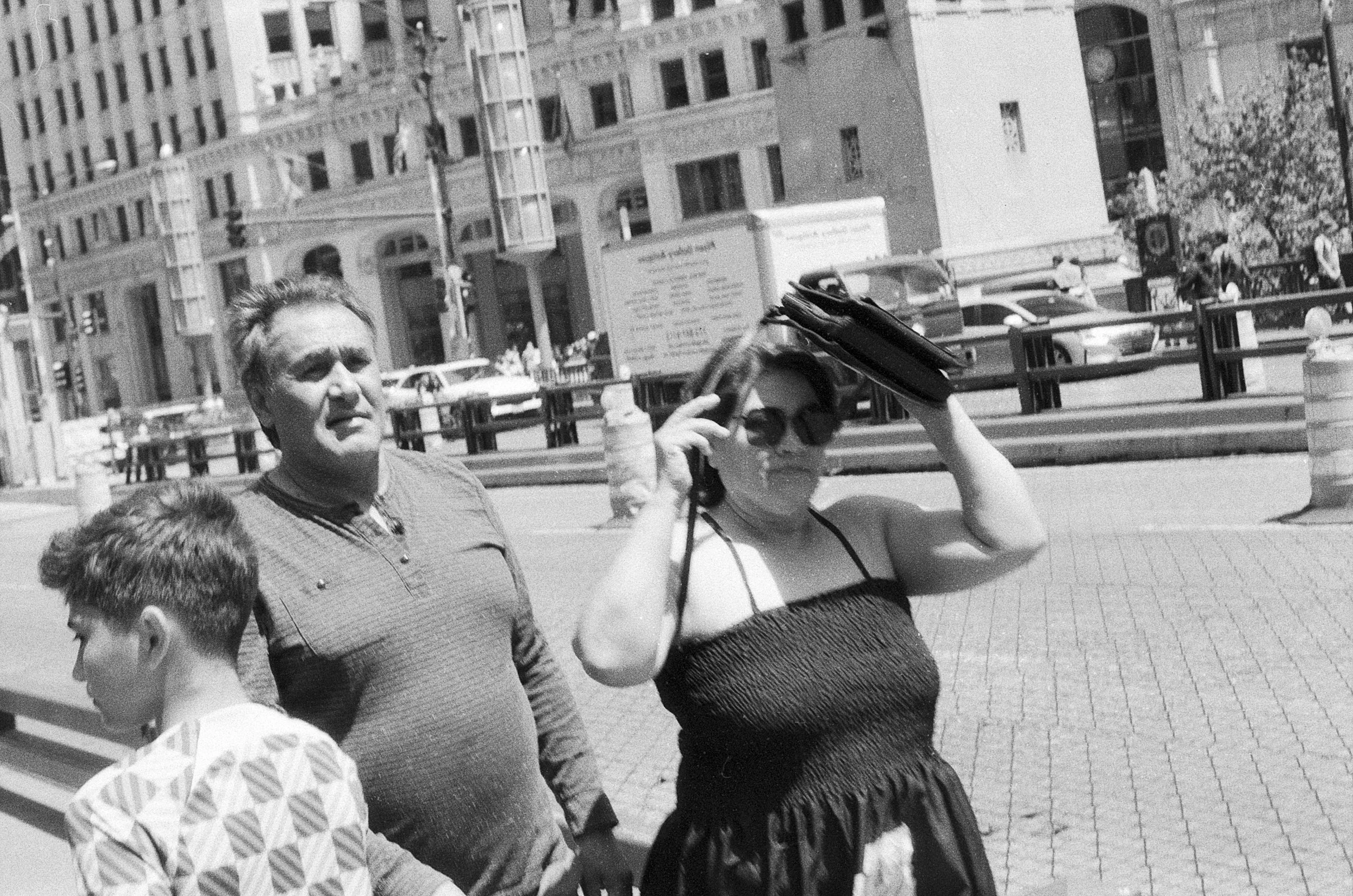 Three people walking on a city street in daylight, with tall buildings and cars in the background. The woman in the center is adjusting her sunglasses and holding a purse, the man on the left is looking ahead, and the boy on the right is looking down
