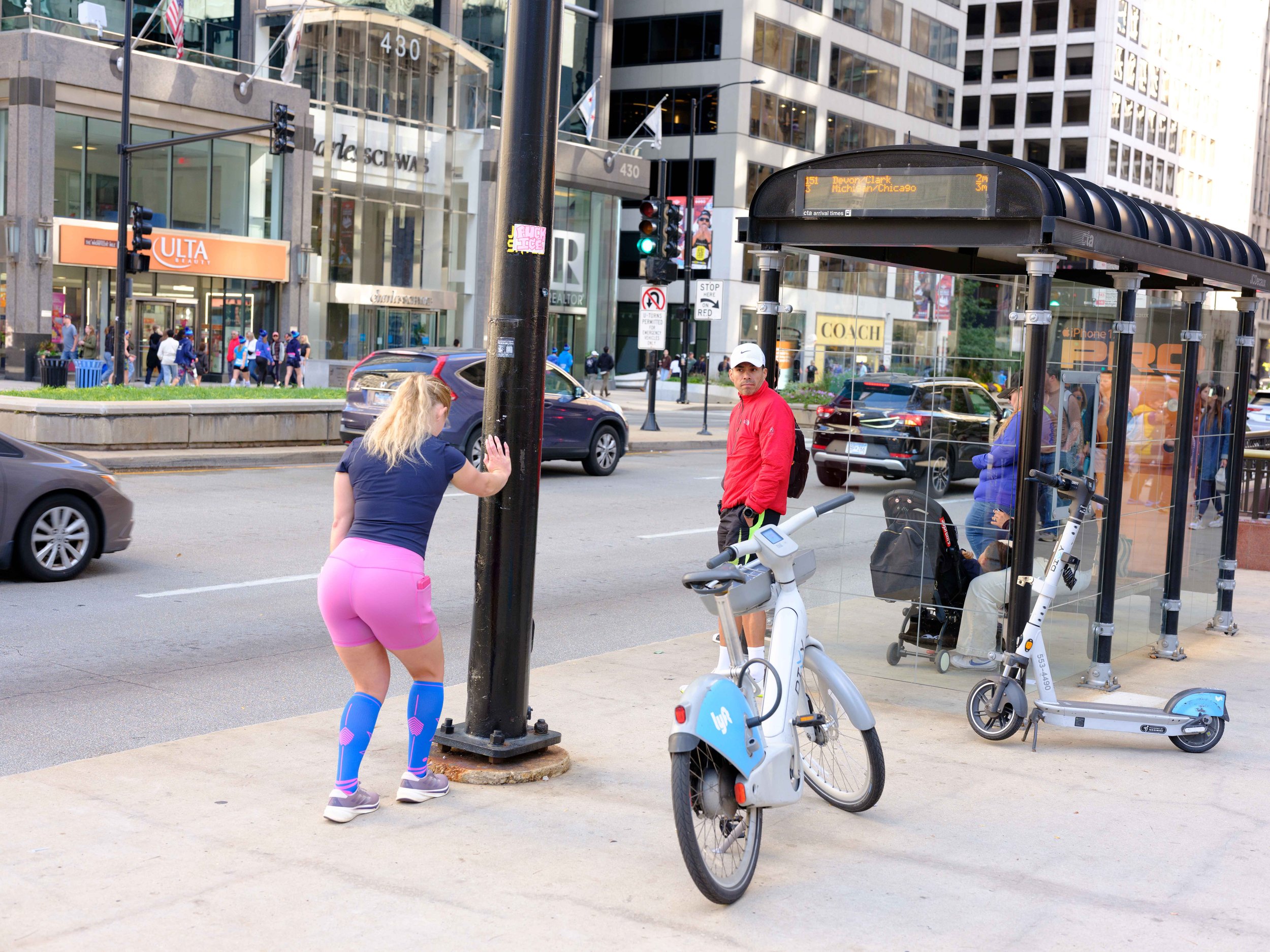 A woman in workout clothes leaning against a street lamp while a man in a red jacket stands near a bus stop in an urban area with tall buildings and traffic.