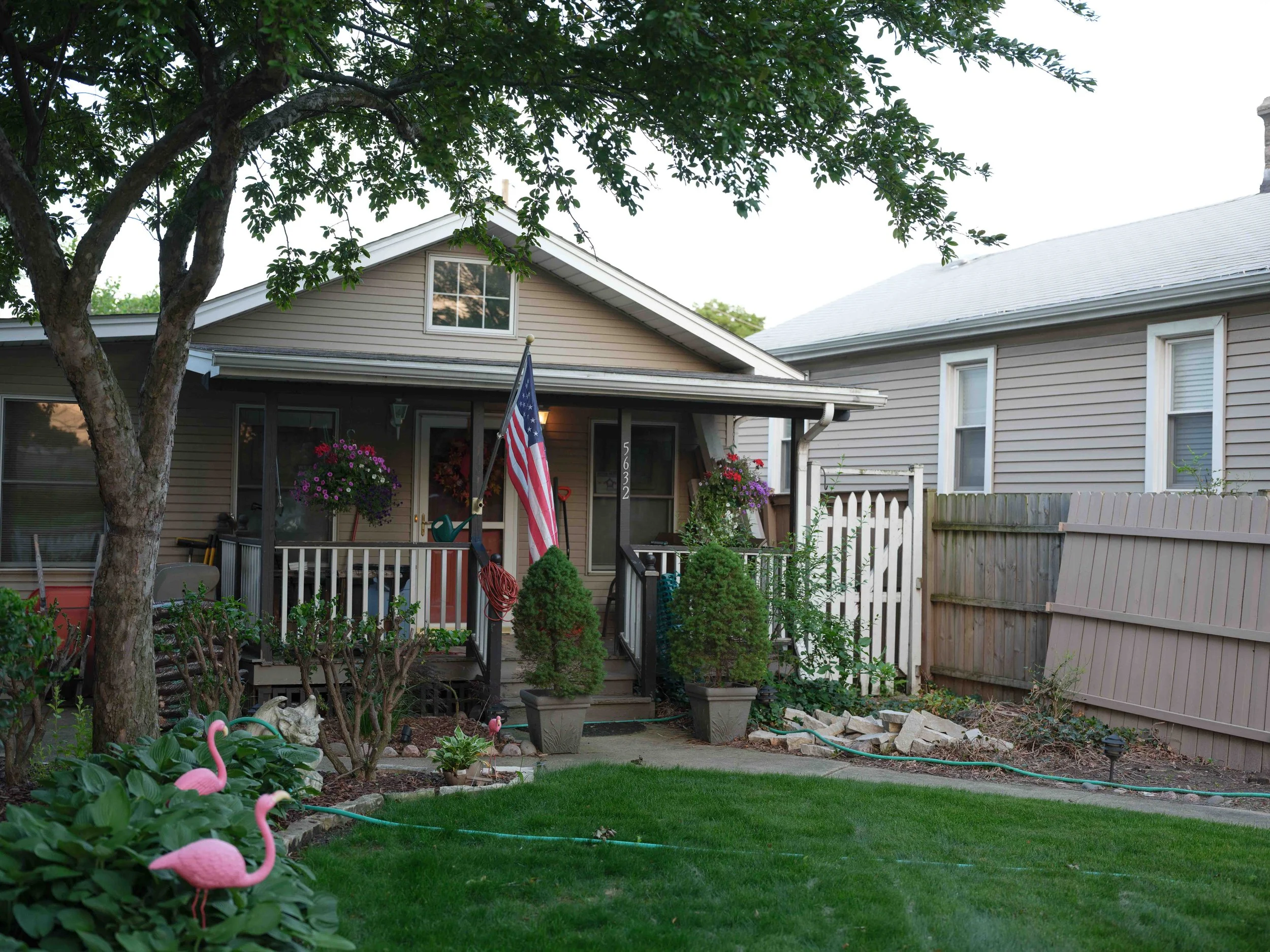 A house with a small front porch decorated with hanging flower baskets, an American flag, and potted plants. The front yard has a green lawn, pink flamingo lawn ornaments, and a garden with shrubs and flowers. A cat is resting near the bushes, and a 