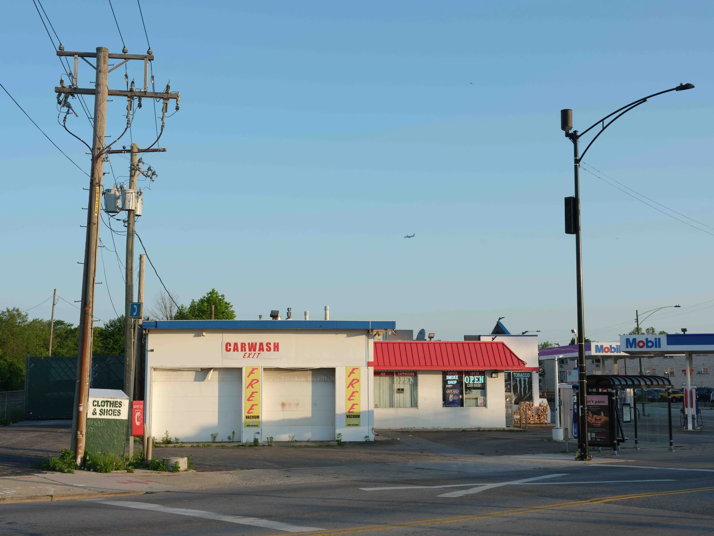 A small convenience store with a car wash, gas station, and bus shelter, with a clear blue sky and an airplane flying overhead.