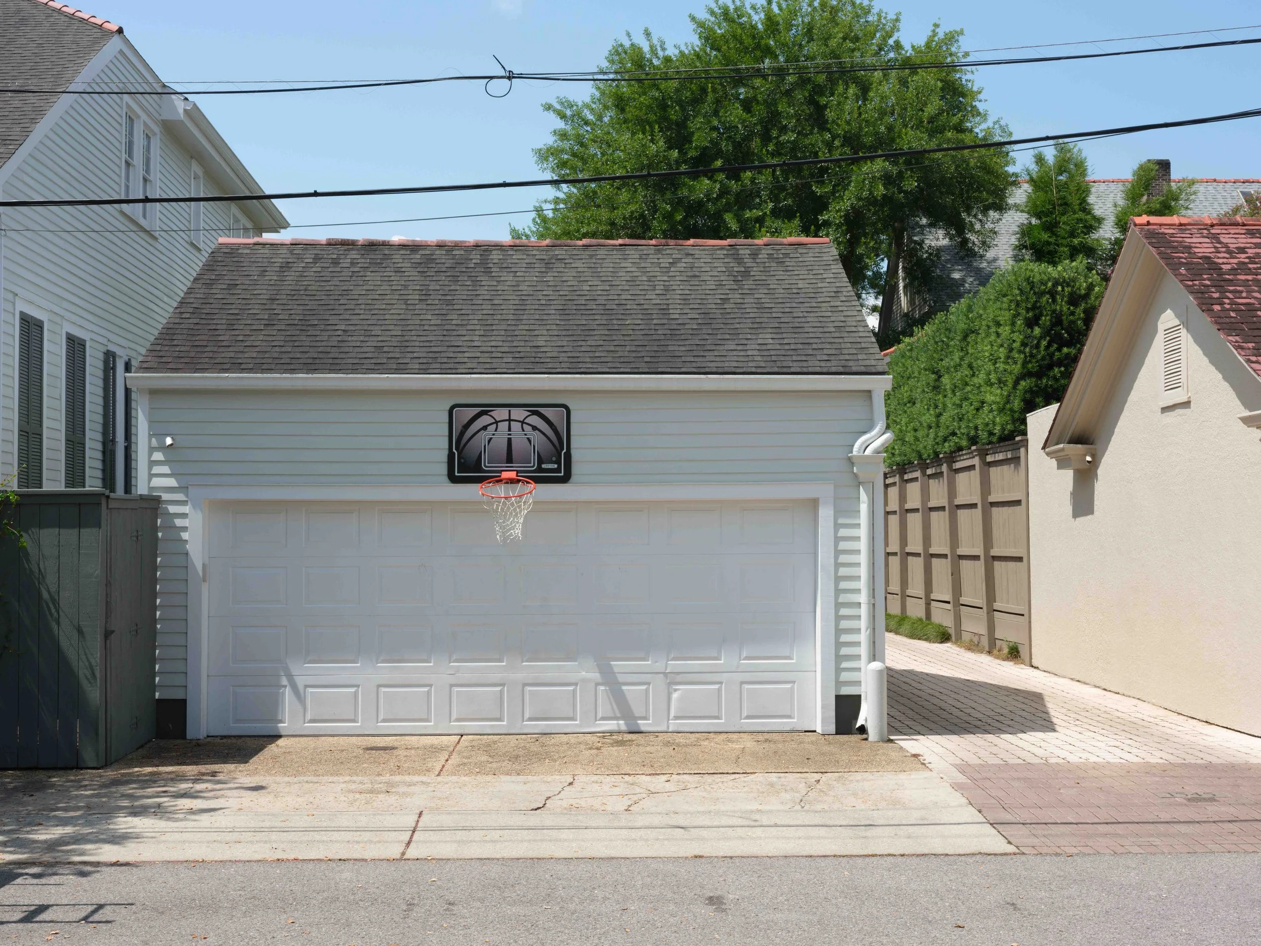 A small white garage with a basketball hoop attached above the door, situated in a residential neighborhood with neighboring houses, trees, and power lines visible in the background.