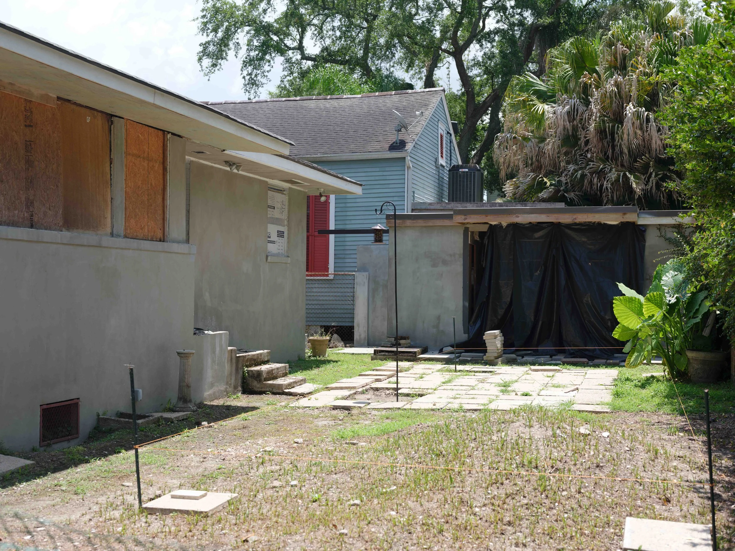 Backyard with two small buildings, one under construction with plywood and stucco, another covered with black tarp, patio stones, grass patch, and plants, with trees and neighboring houses in the background.