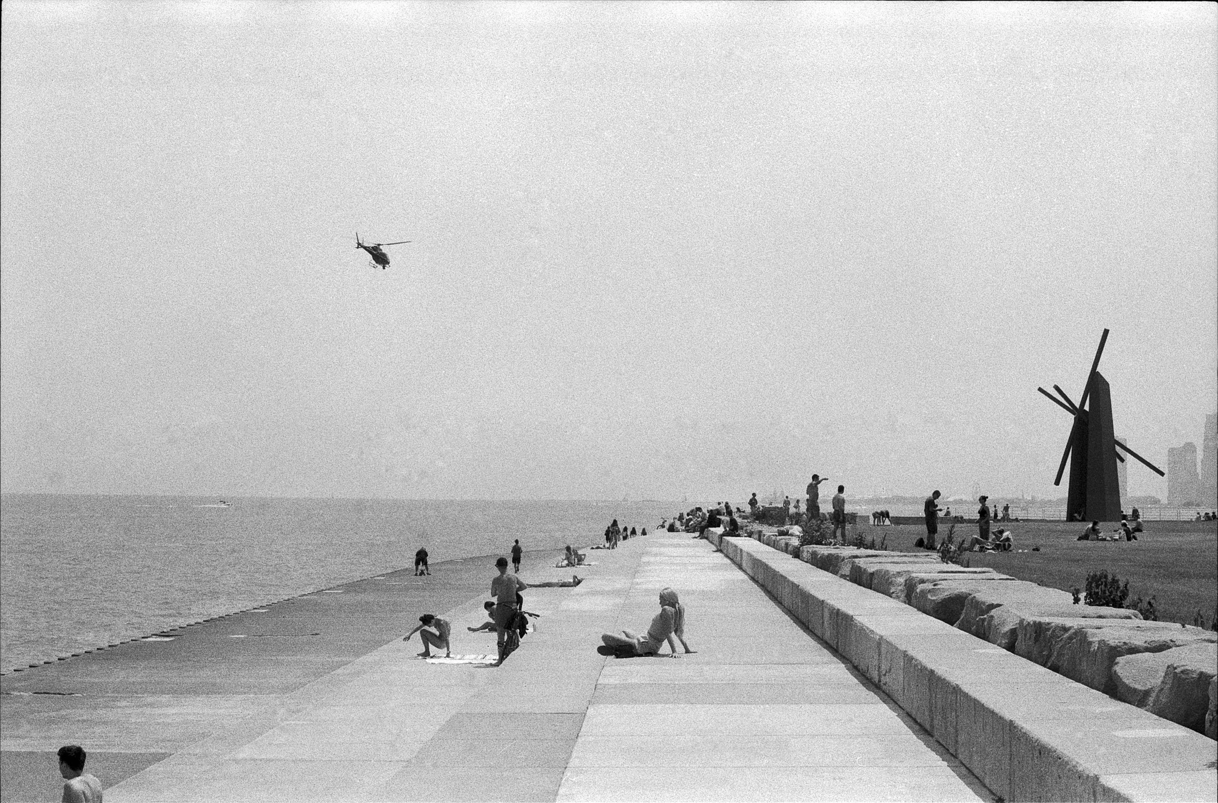 People relaxing along a waterfront promenade with a large modern sculpture and a windmill in the background, and a helicopter flying in the sky.