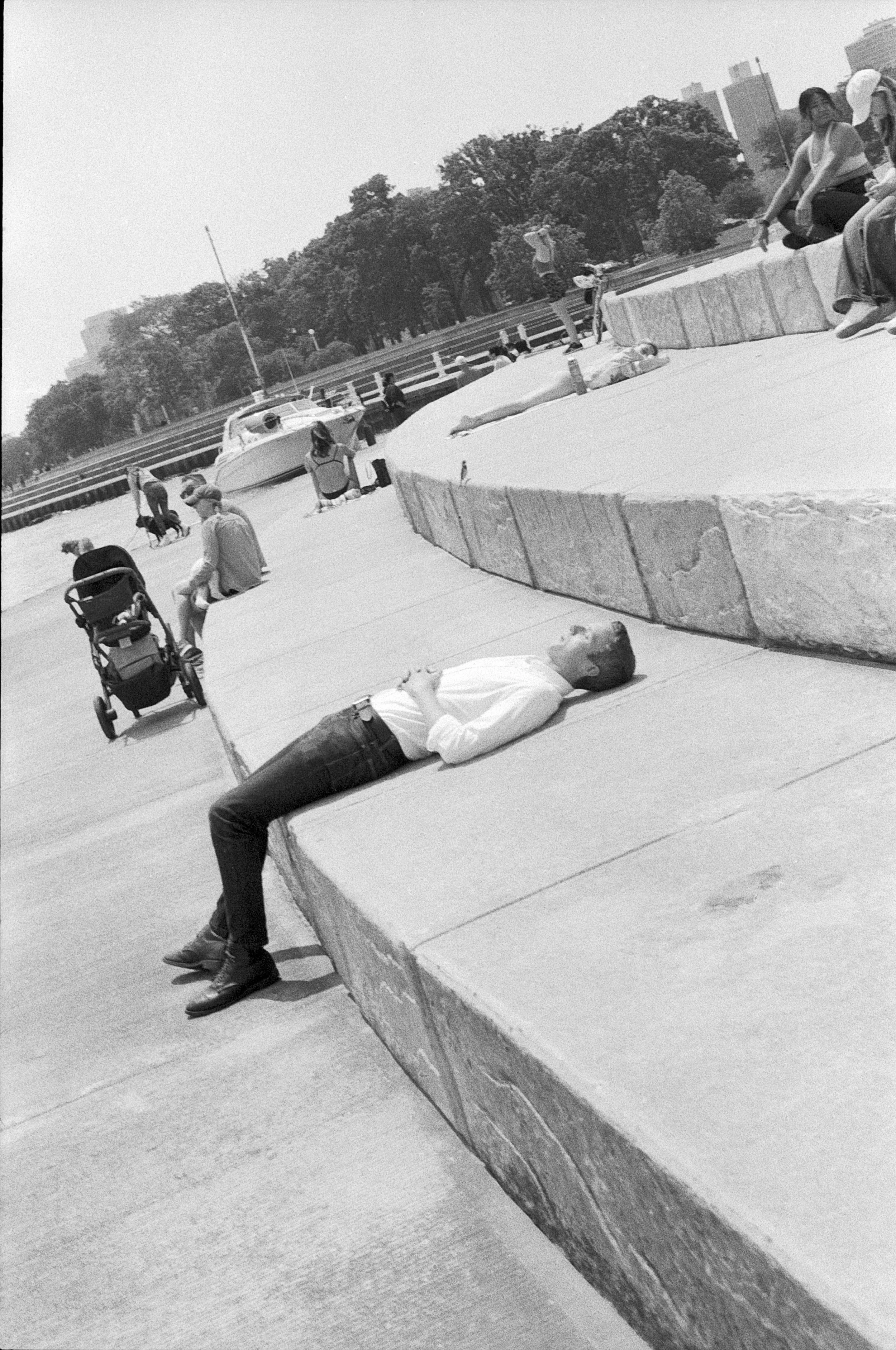 A man lying on a large stone ledge by the water with his eyes closed, wearing a white shirt and dark pants, in a park or waterfront area with people walking and sitting, trees, and a boat in the background.