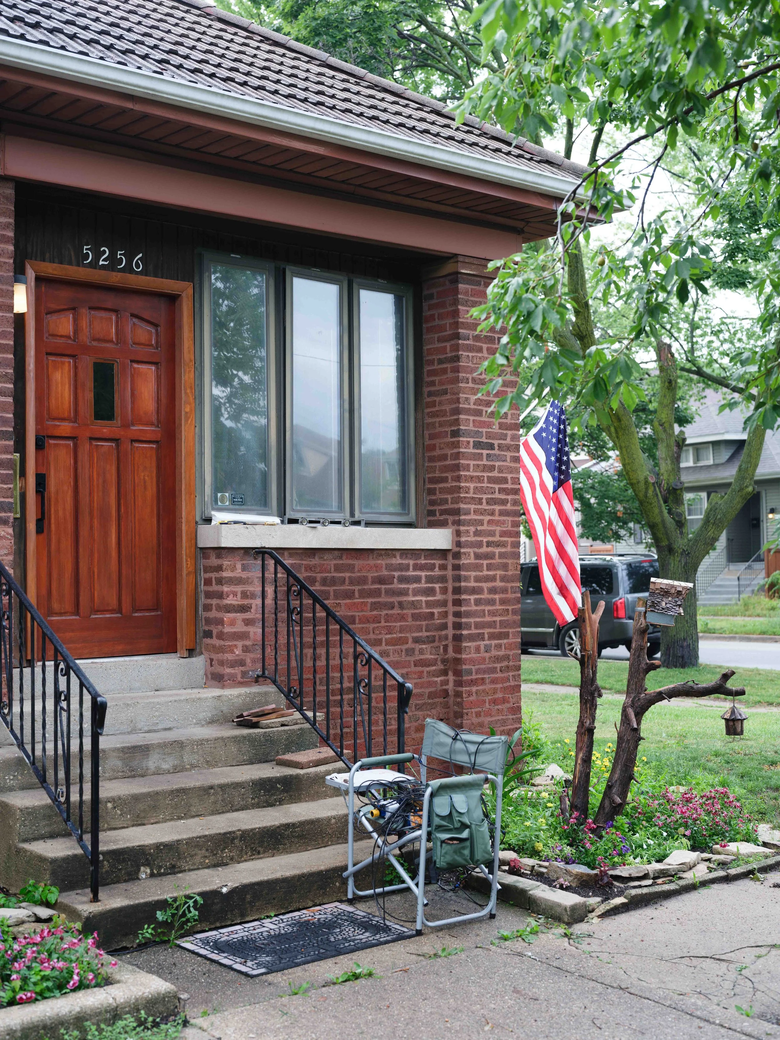 A brick house with a wooden front door, a large window, and stairs leading up to the entrance. There is an American flag attached to a tree on the front yard, with some pink flowers and green plants nearby. A small folding chair with cables and a bag