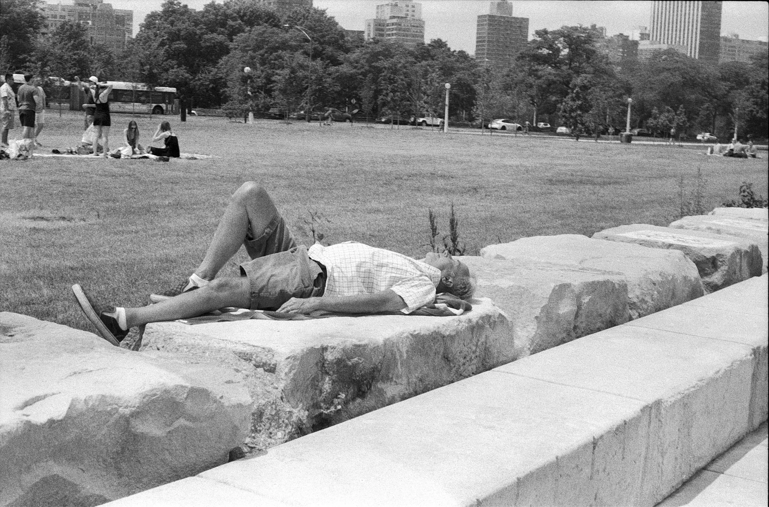 An elderly man lying on a blanket on large rocks in a park, resting with eyes closed, wearing a plaid shirt, shorts, and sneakers. In the background, people are sitting or standing on the grass, with tall buildings and trees visible.