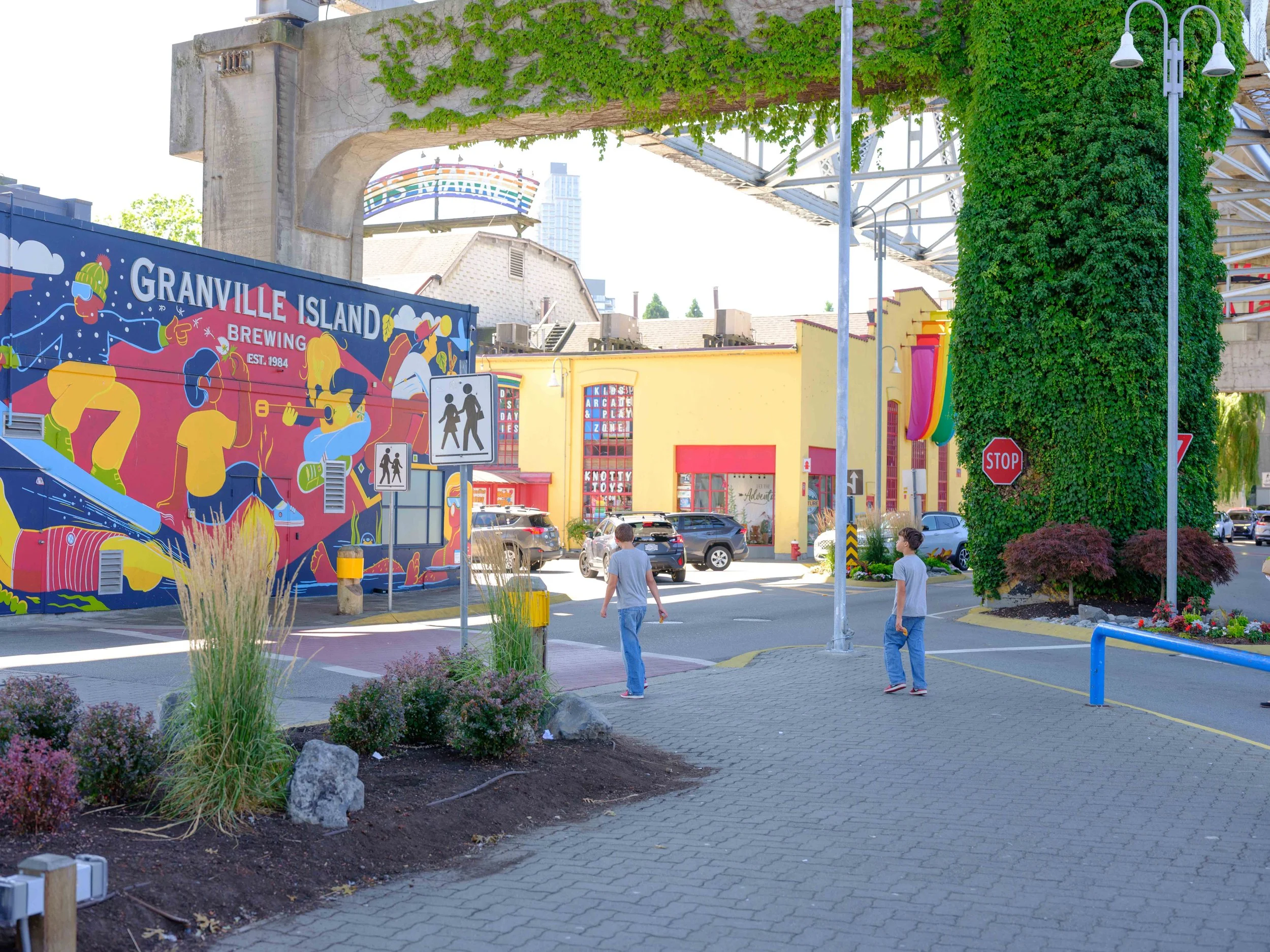 Street scene with a colorful mural on a building, pedestrians, stop sign, streetlights, and lush greenery
