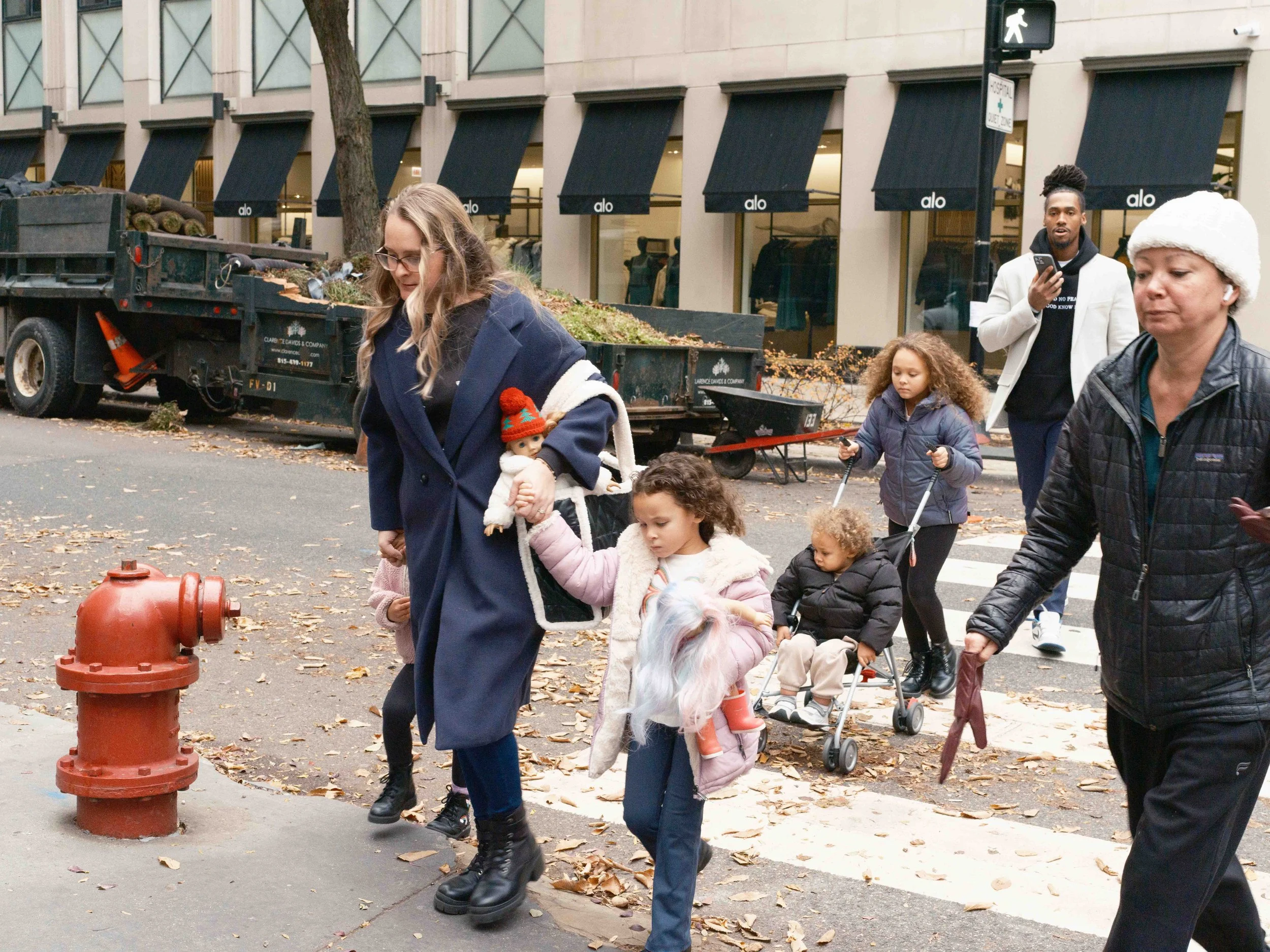 A diverse group of people crossing a city street during the fall, some walking and some in wheelchairs, with fallen leaves on the ground.
