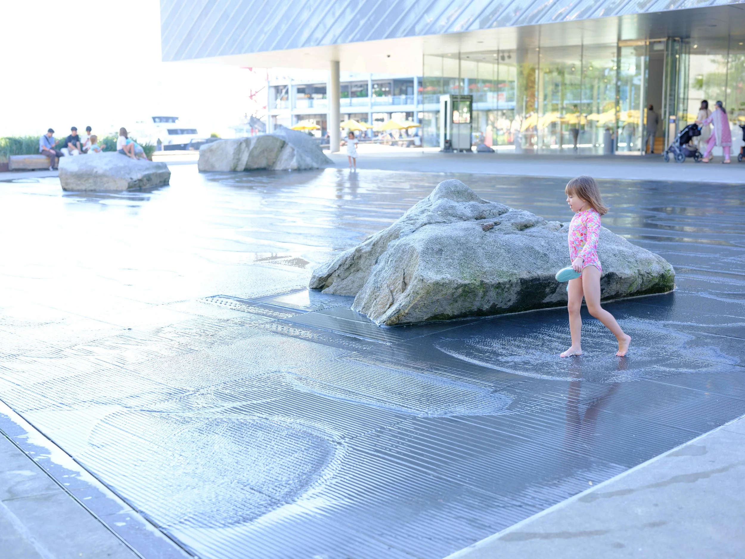 A young girl in a pink floral swimsuit playing in shallow water near large rocks outside a modern building with a glass facade.
