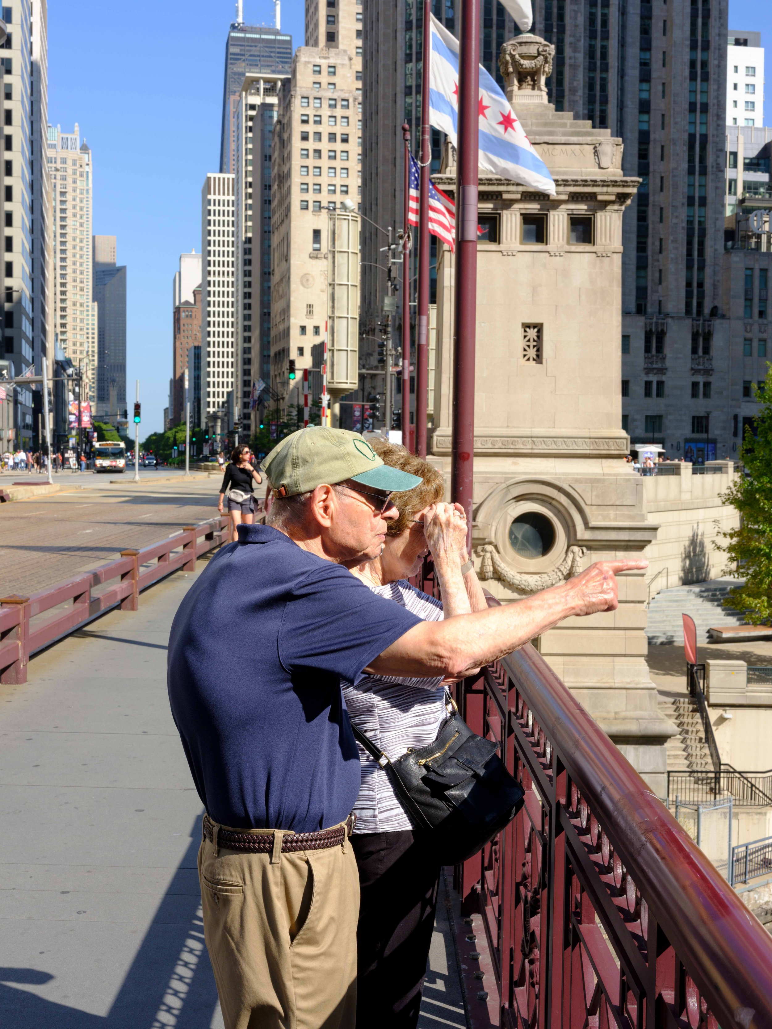 Two elderly people standing on a city bridge, looking and pointing at something in the distance with a busy cityscape behind them. There are American flags and flags with a red star, and tall buildings in the background.