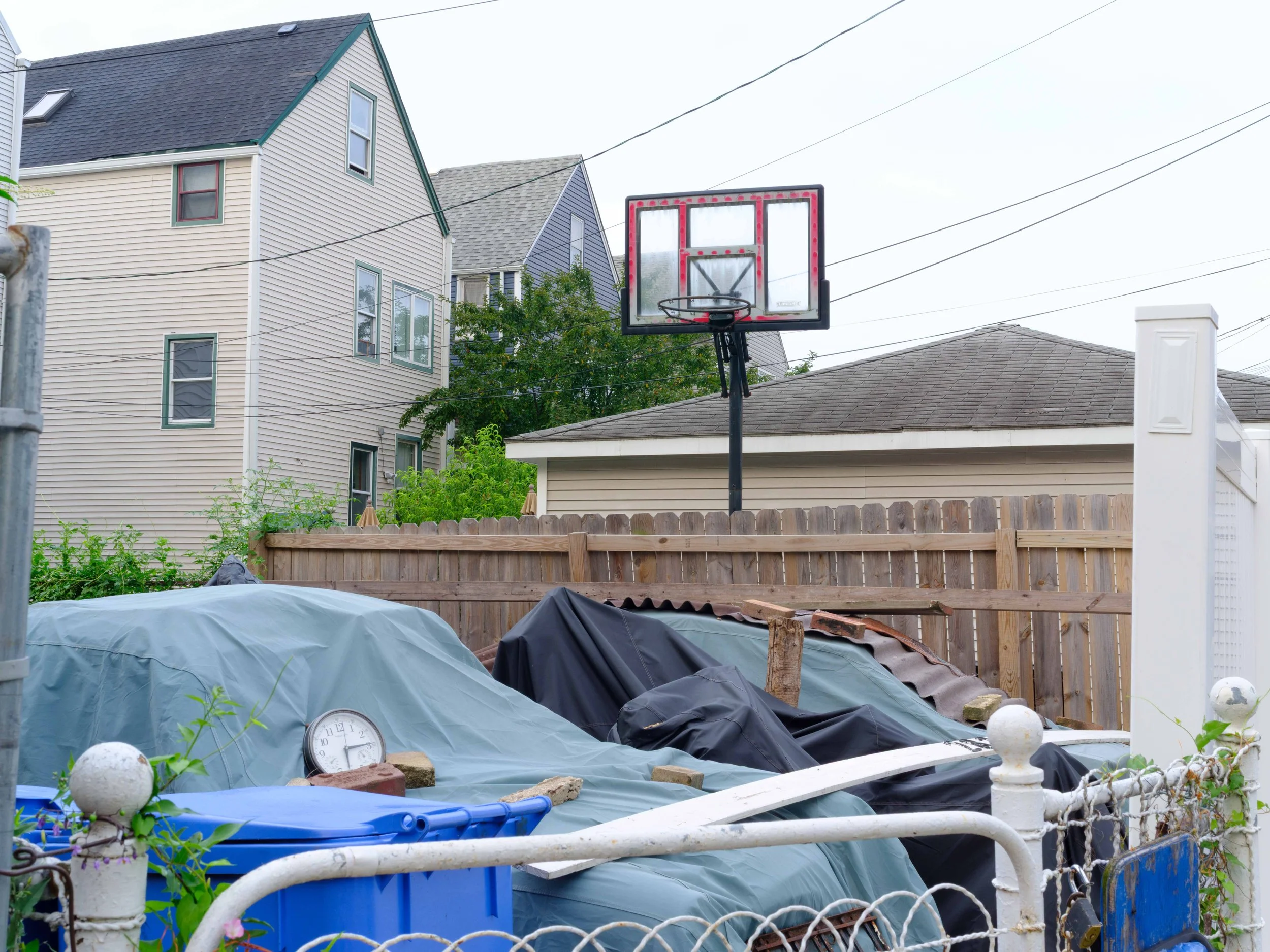 Backyard with a basketball hoop, covered items, and a white fence, residential houses in the background.