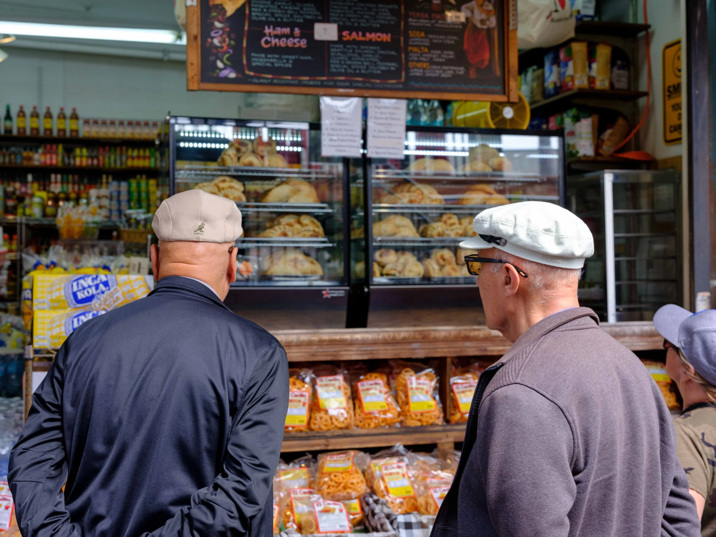 Three elderly men wearing caps and jackets are looking at baked goods displayed at a market or bakery stall, with shelves of packaged snacks and a chalkboard menu in the background.