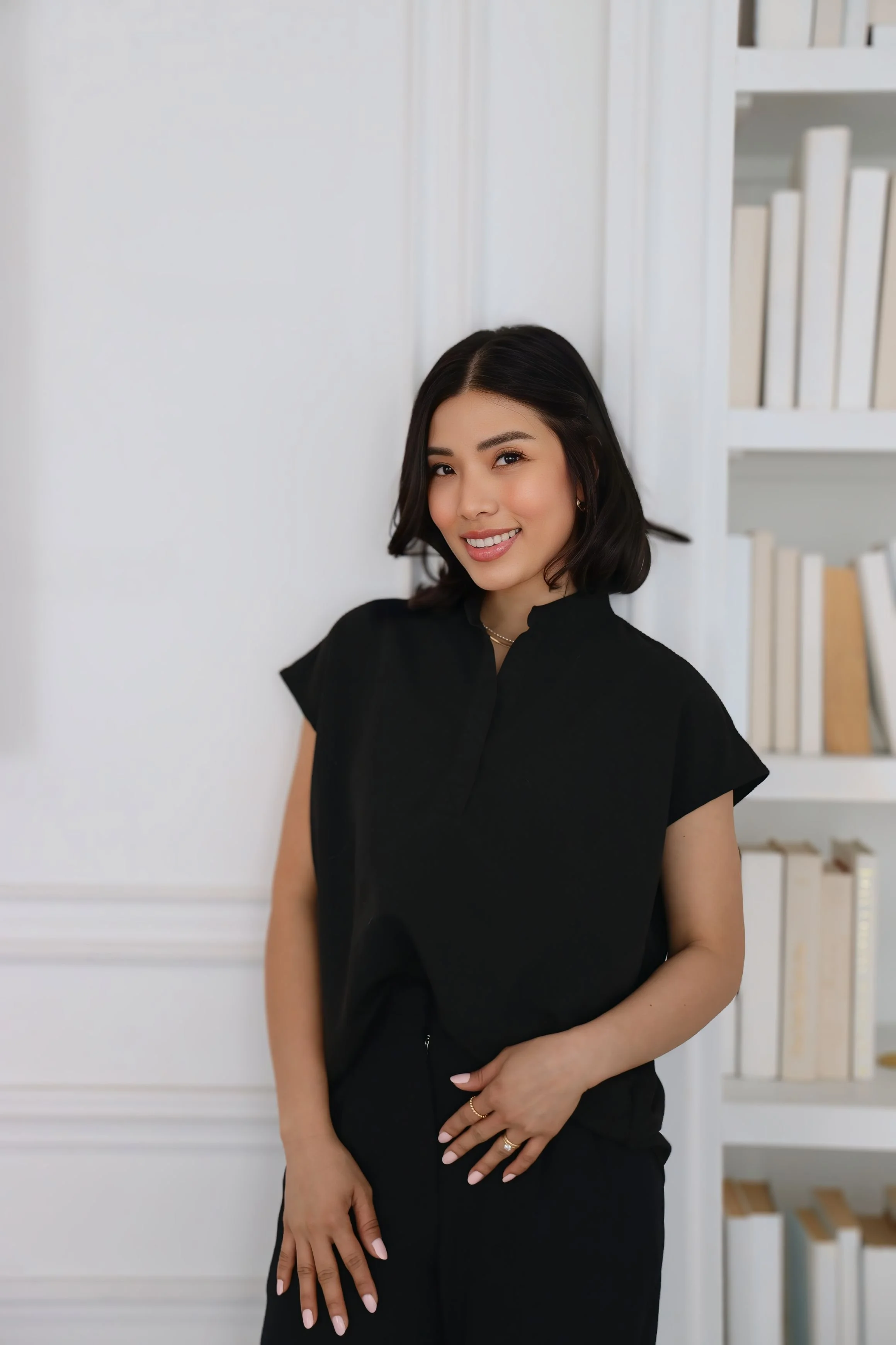 A woman sitting on a cream-colored couch in a minimalist living room, smiling at the camera. Behind her, a white fireplace mantel with decorative vases, a mirror, and abstract artwork.