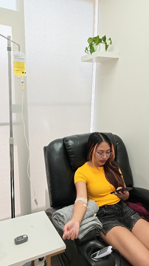 A young woman with glasses and long hair, wearing a yellow shirt and black shorts, sitting in a black recliner chair donating blood at a medical facility. She is looking at her phone, with an IV connected to her arm and a folded gray towel resting on her lap.