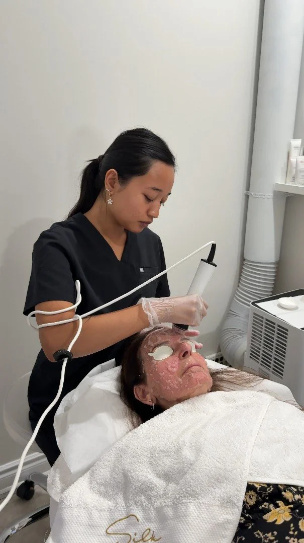 A woman receiving a facial treatment in a spa or clinic setting, with a skincare specialist applying a facial device to her face, while she lies on a bed covered with a towel.