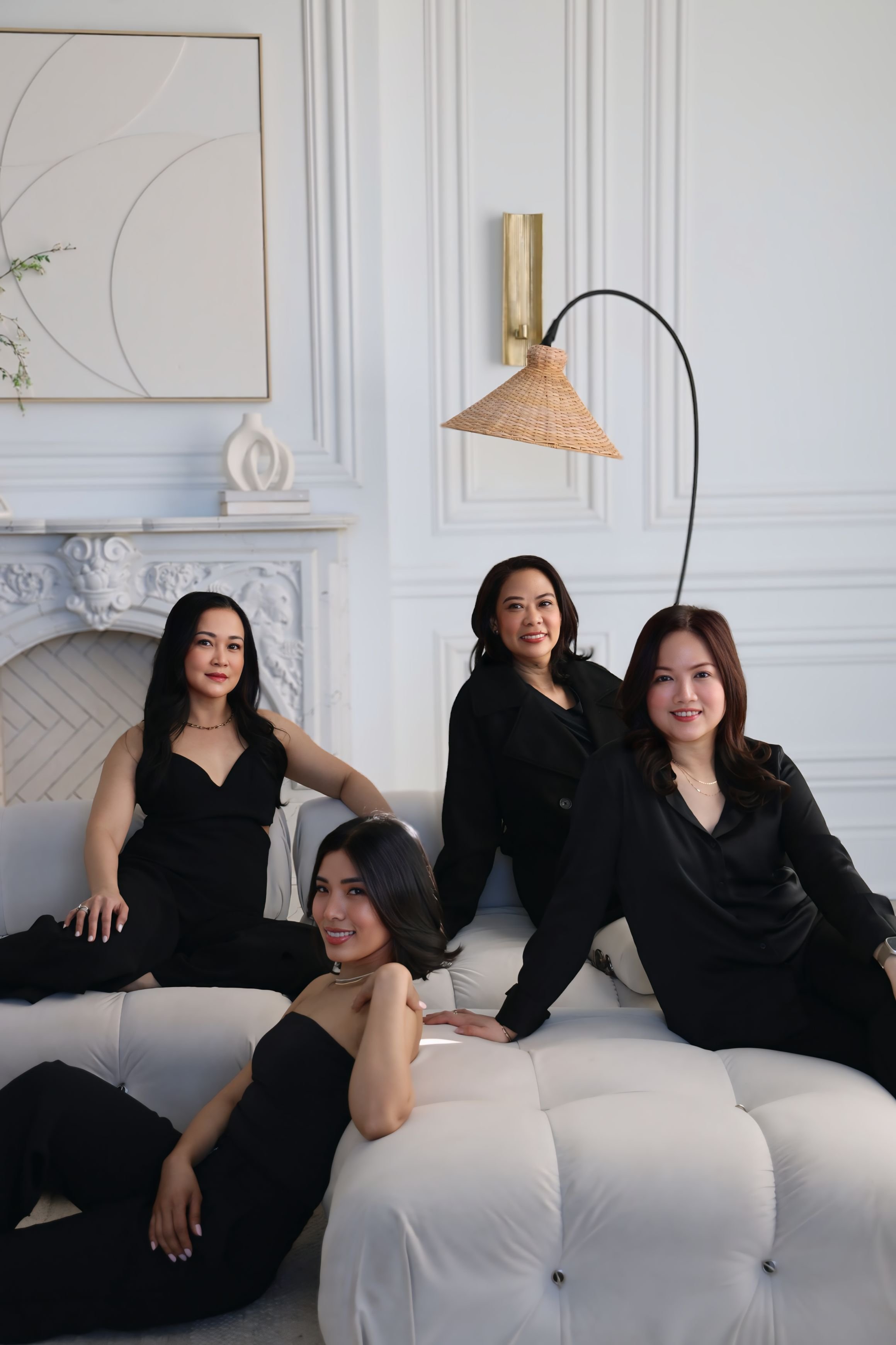 Group of five women sitting and standing on a white couch in an elegant room with white walls, decorative molding, a gold wall sconce, and modern artwork.