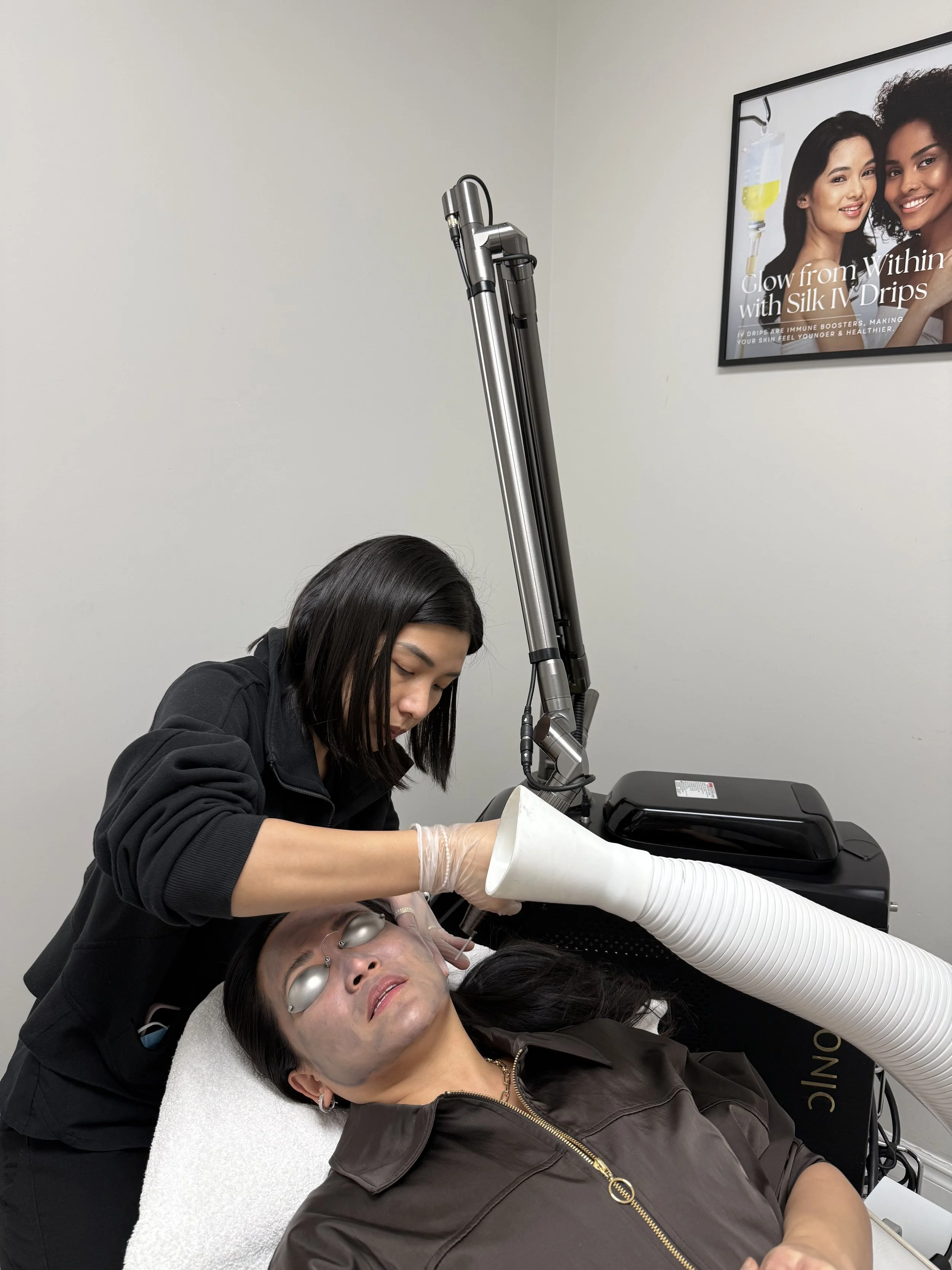 A woman receiving a cosmetic or medical treatment on her face while lying down, with a technician performing the procedure using specialized equipment, in a clinical setting with a framed promotional poster on the wall.