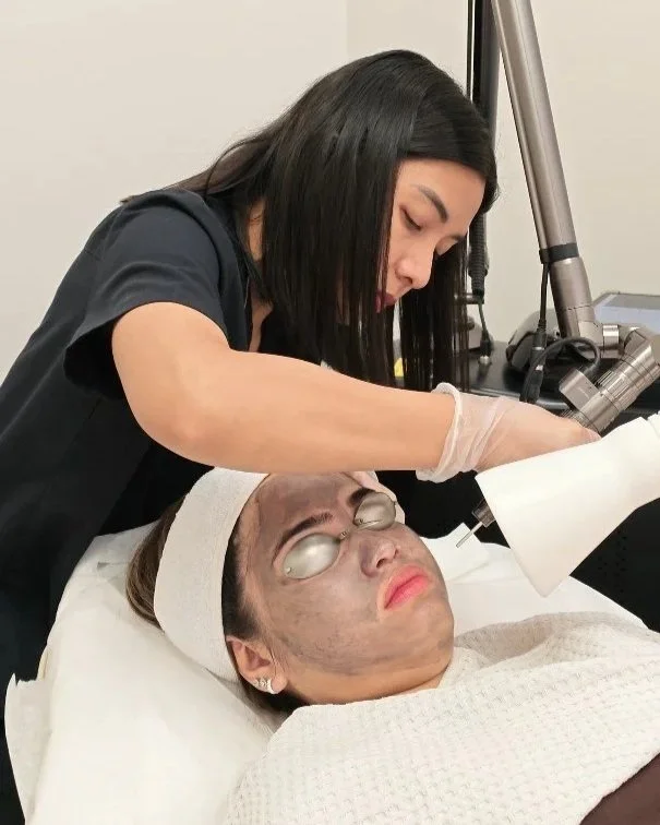 A woman with dark hair and a glove on her hand is performing a cosmetic procedure on a woman lying down, who has closed eyes, glasses, and a headband, in a clinical setting with medical equipment nearby.