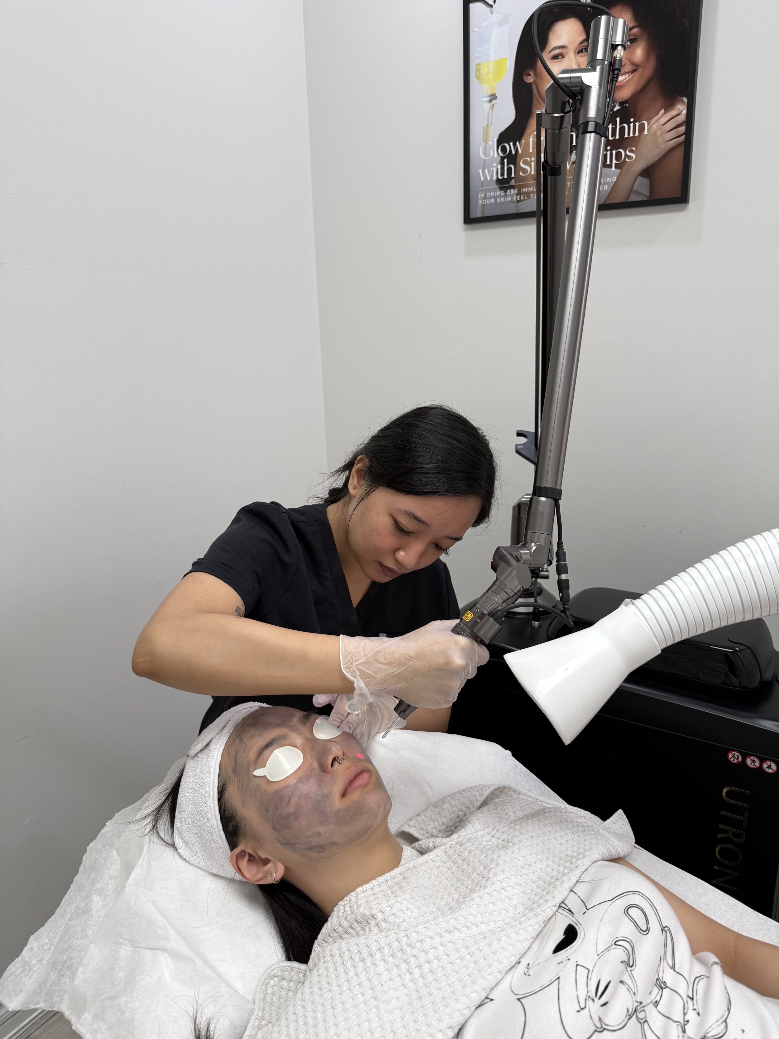 A woman receiving a facial treatment on her face from a skincare specialist in a clinic or spa setting, with her eyes covered by eye patches and a white towel wrapped around her head.